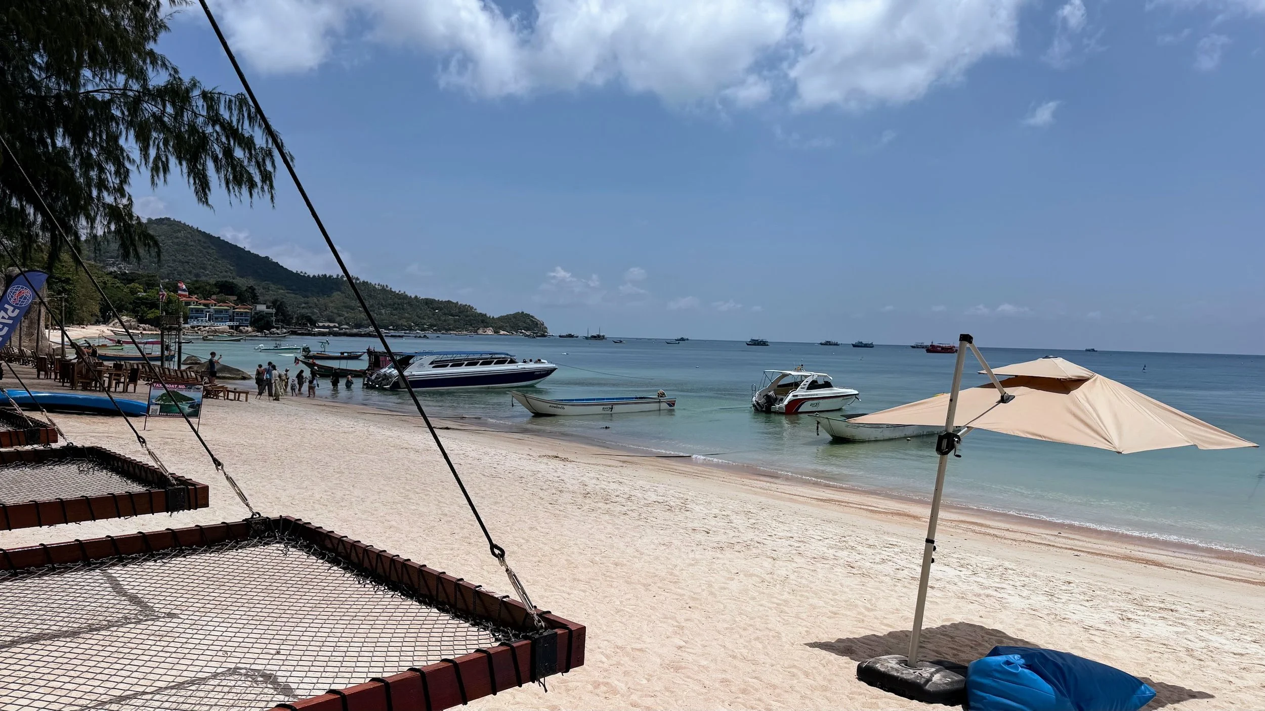 Wooden frames with nets hang over a Sairee Beach in Koh Tao, steps from the ocean, with boats on the water in a bay, and a green hilly peninsula in the background.
