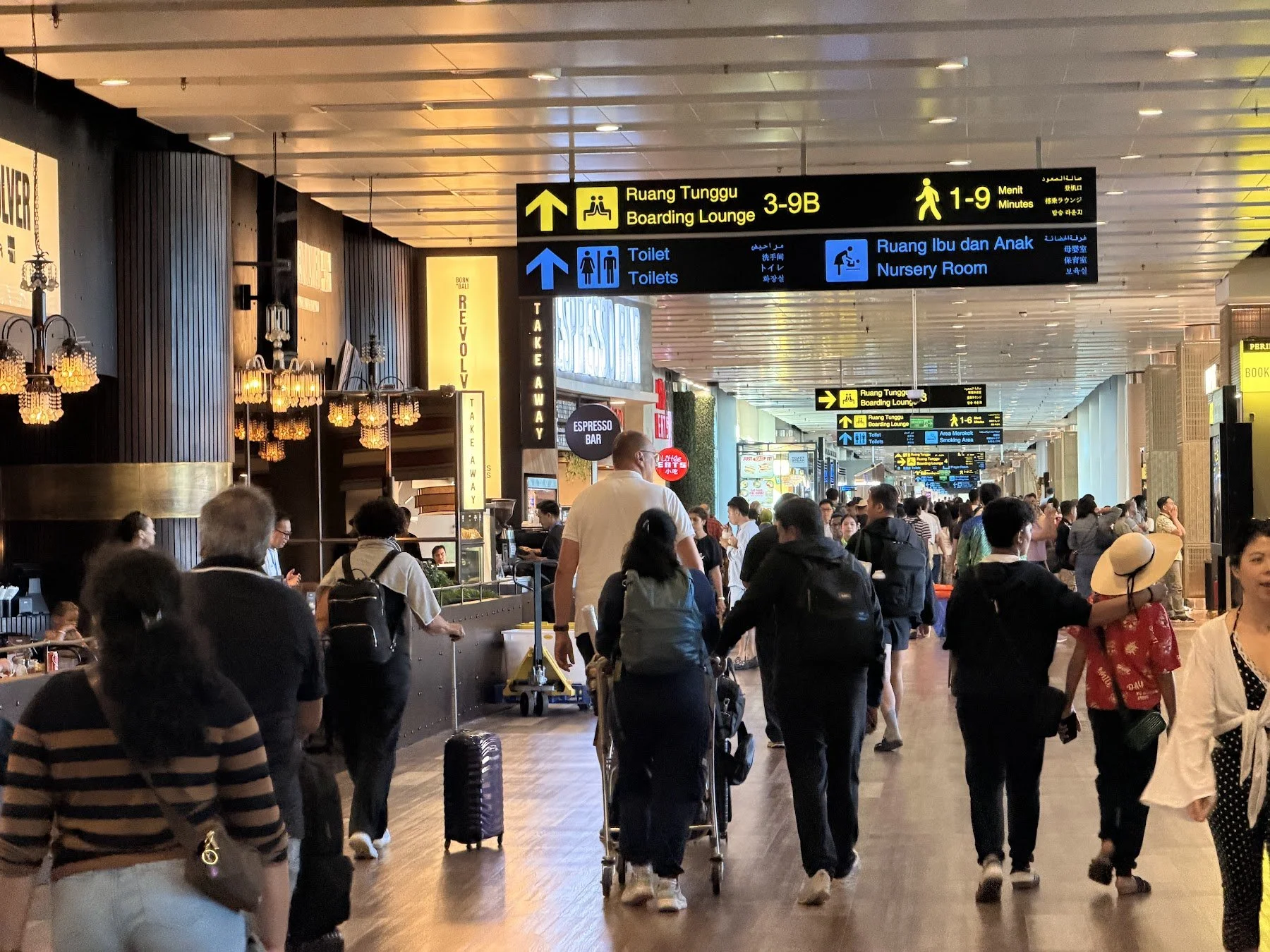 Inside a busy airport departures hall, with restaurants lining the left side, many passengers walking the hall with luggage, gates to the right, and signage overhead.