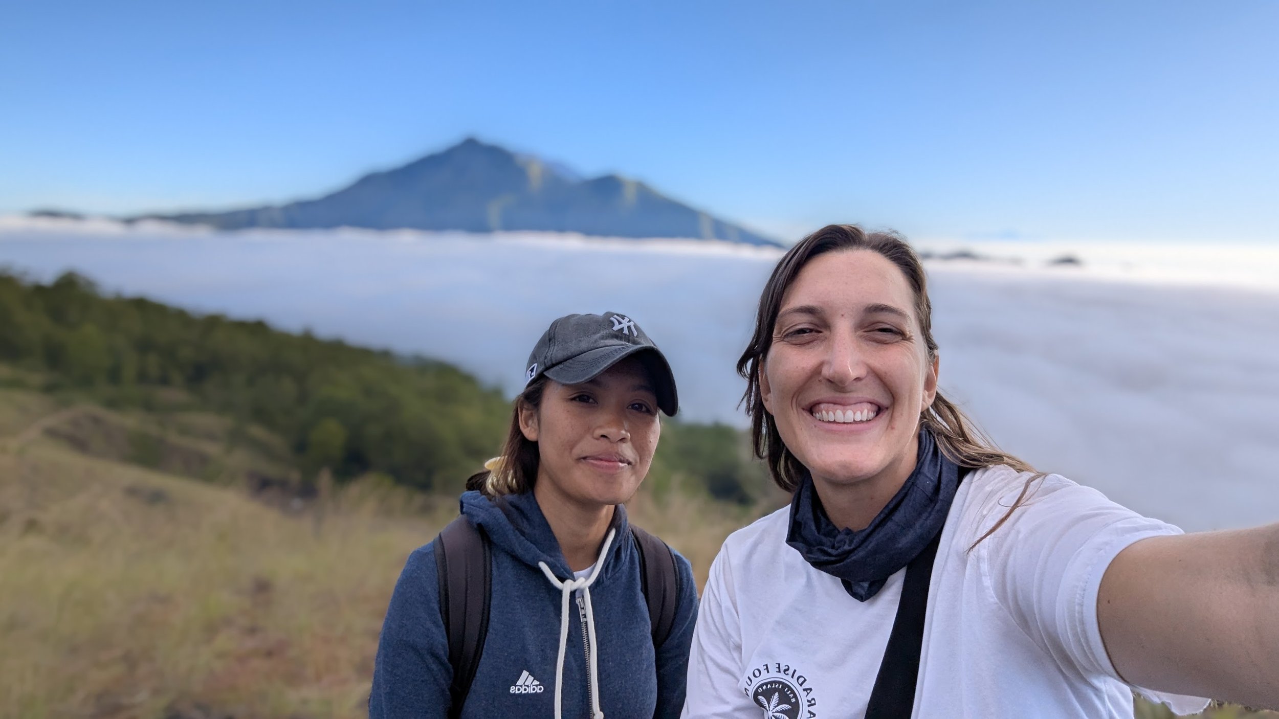 Lynne and hiking guide Kiki take a selfie from Mount Batur in Bali with a sheet of clouds below and mountain in the background