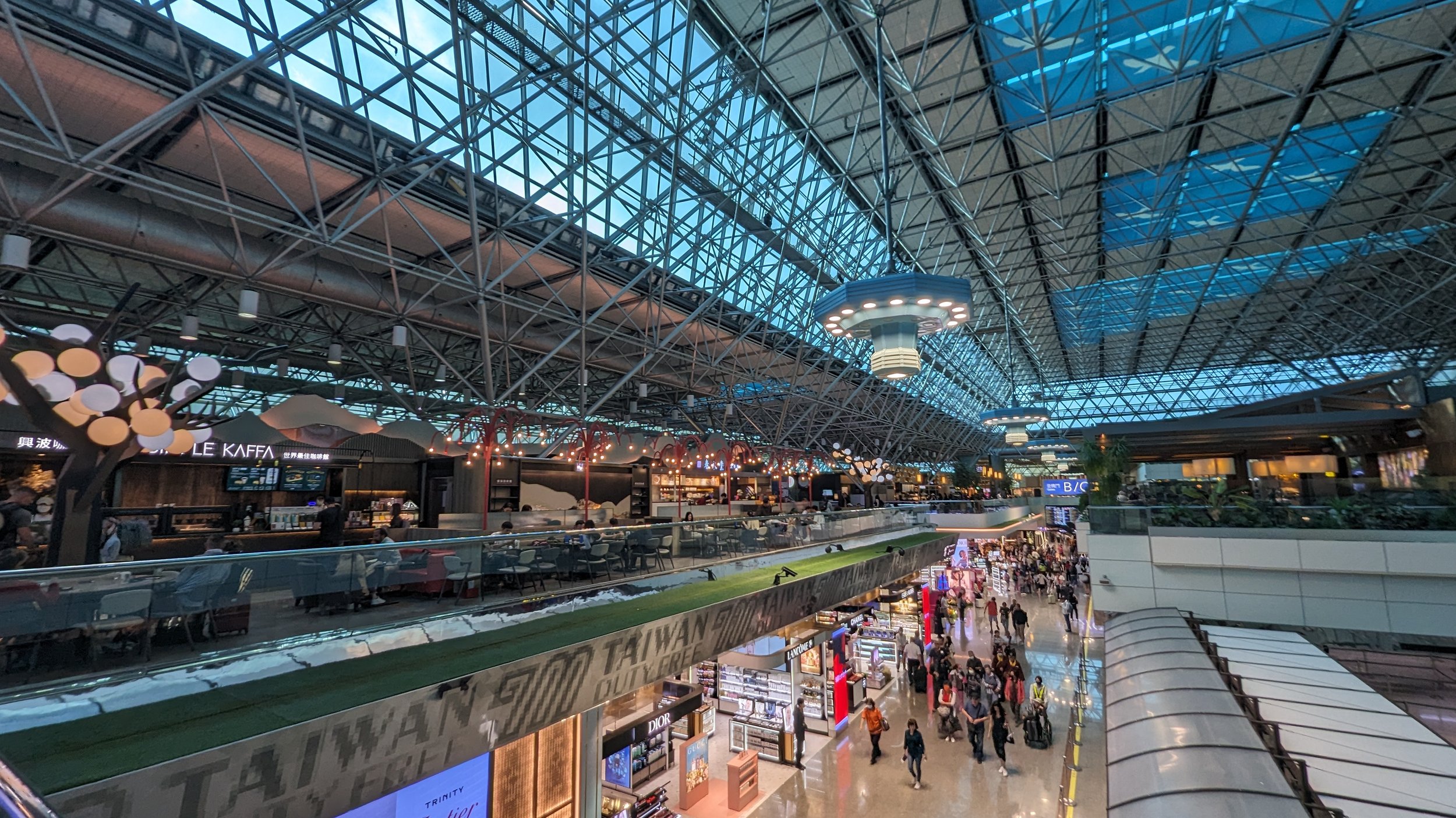 A view of Taiwan Taoyuan International Airport from a mezzanine, with passengers walking alongside shops below, restaurants on the mezzanine level, and large windows overhead.