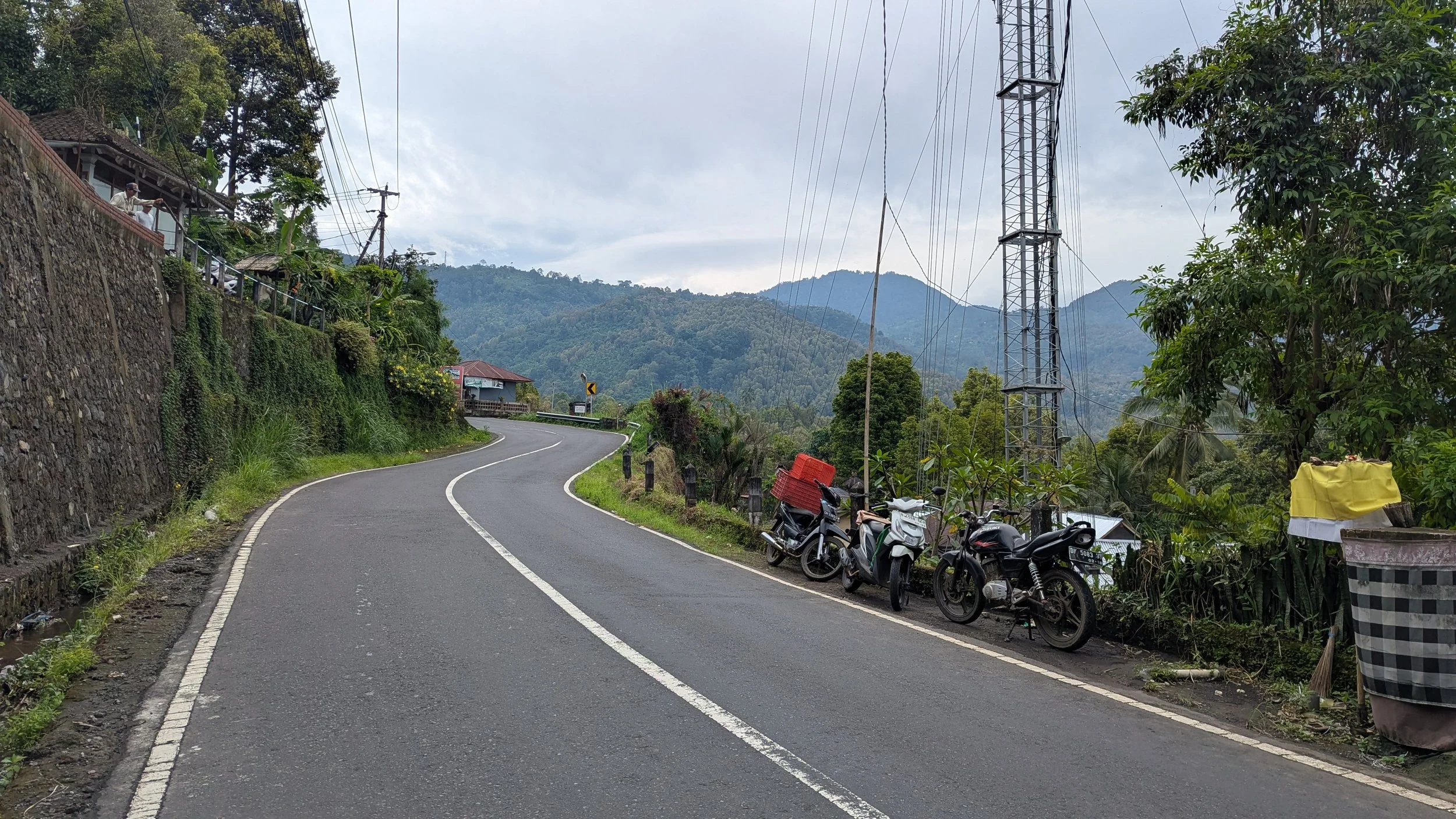 A concrete road in Bali winds through rolling green hills, with a stone wall reinforcing one side and motorbikes lining the other side.