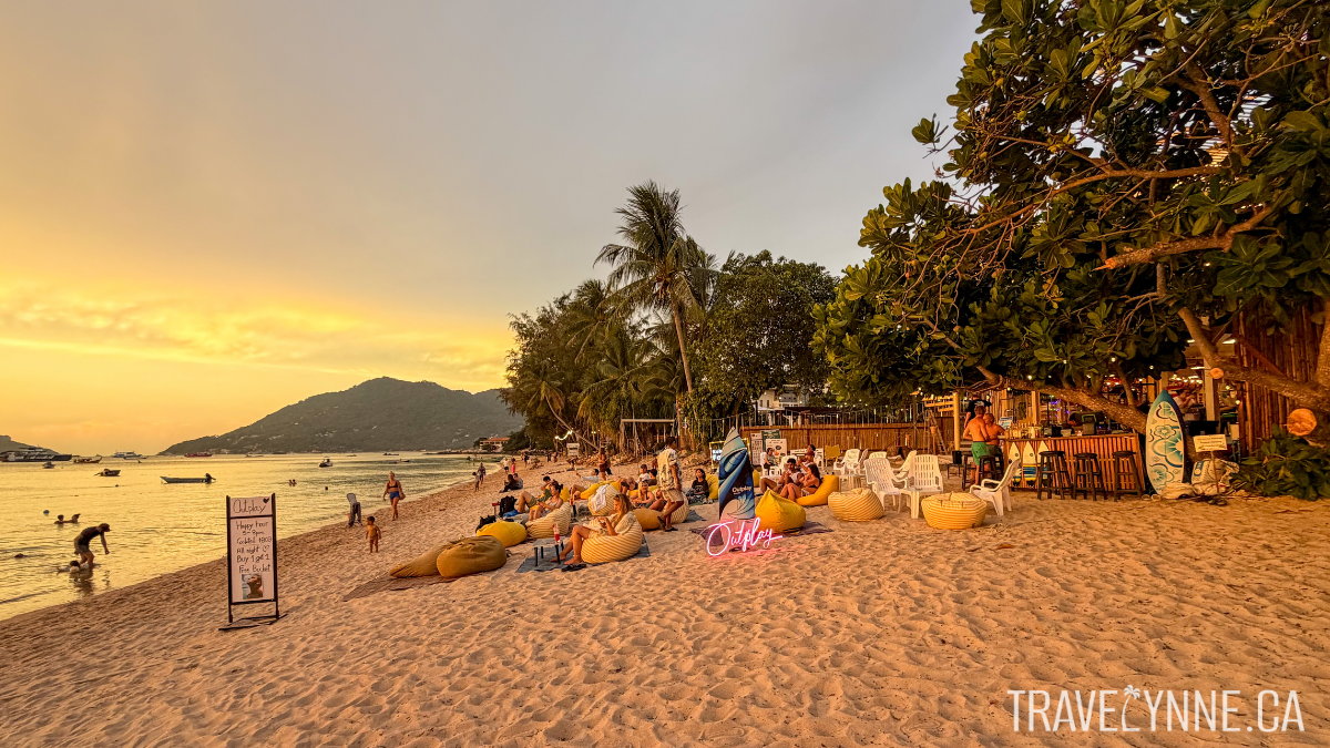 Yellow bean bags line a beige sandy beach during the golden hour while people watch the sunset.