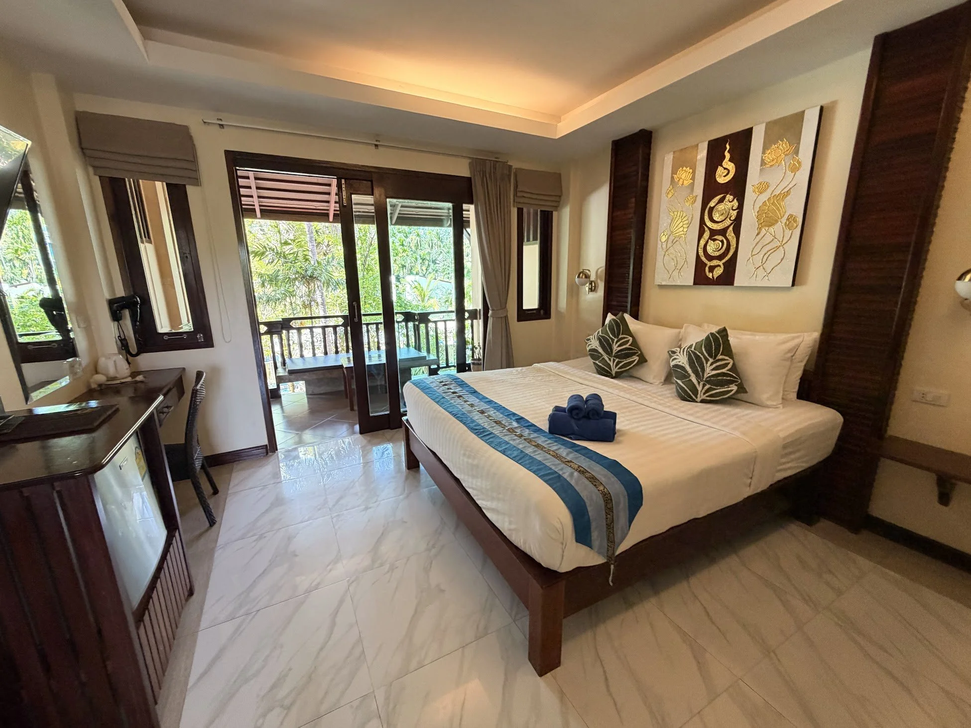 A hotel room at Wind Beach Resort, Koh Tao, Thailand, with a queen bed, a desk, chair and mini fridge, marble tiled floors, glass patio doors to a balcony, and greenery outside.