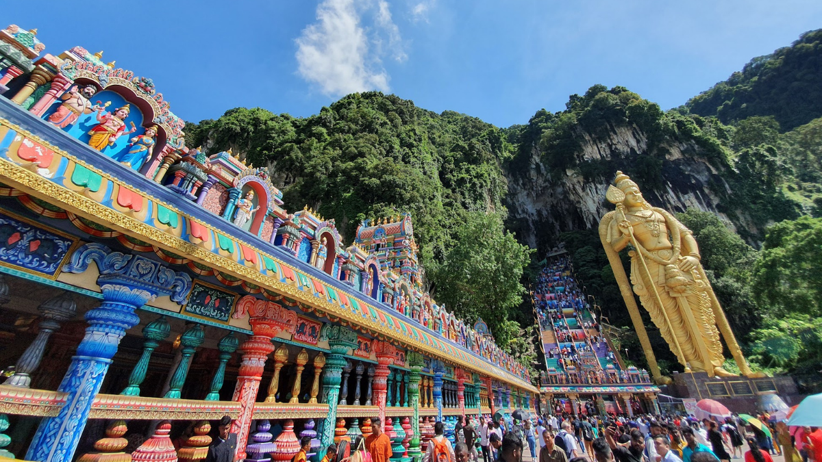 A hindu temple called Batu Caves is painted in bright multicoloured patterns, with a giant golden Lord Murugan statue and rainbow staircase going up into limestone cliffs, and many visitors walking about