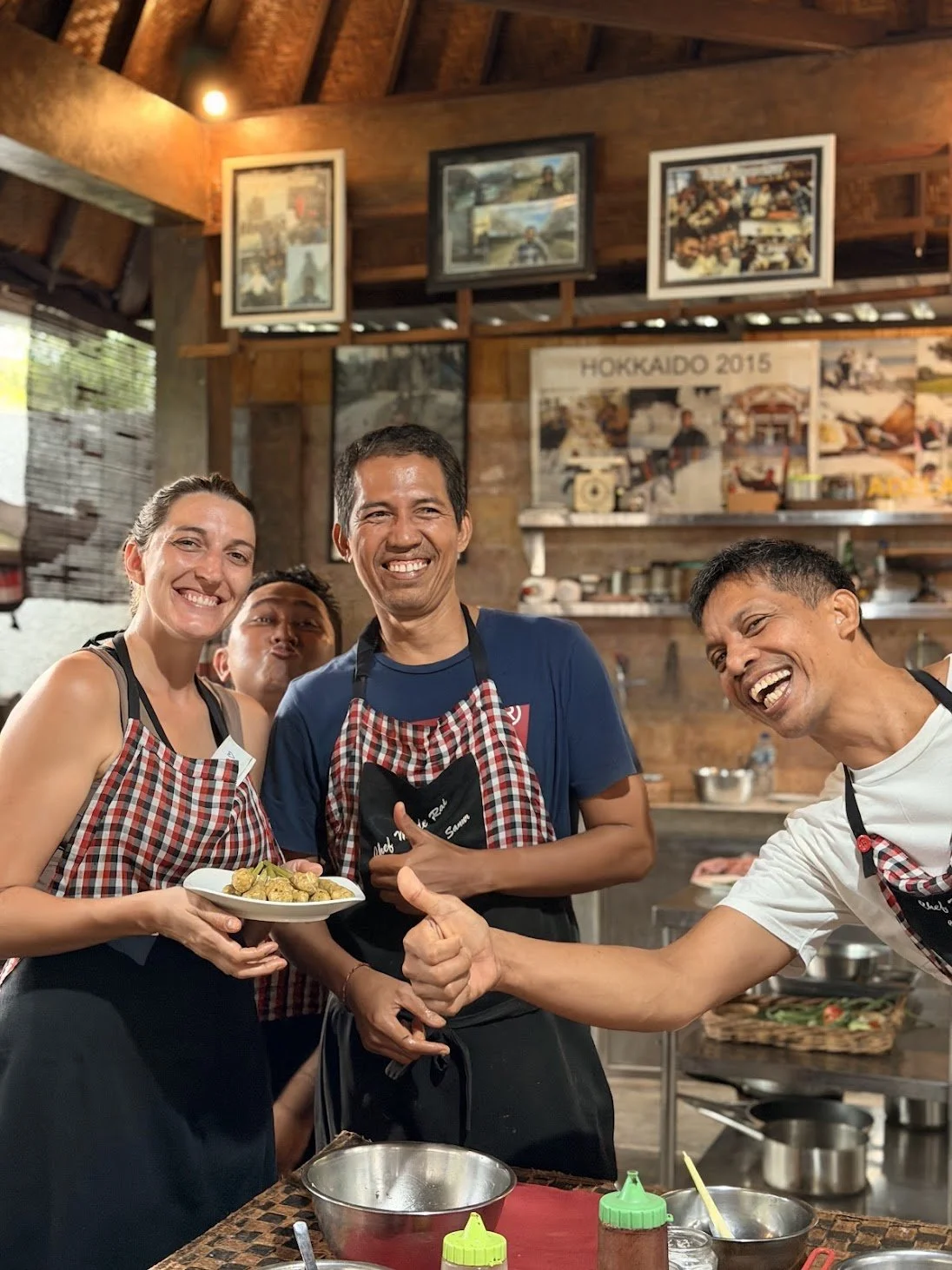 Lynne and 3 Balinese chefs laugh and make silly faces and smiles during a cooking class, holding a plate of satay and wearing aprons