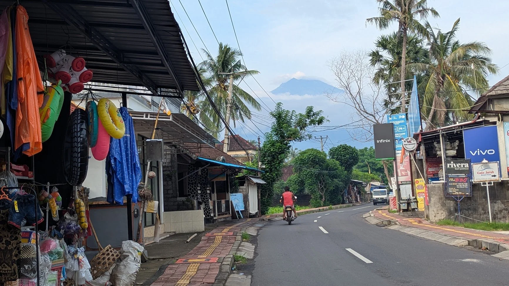Mount Agung volcano stands majestically in view from a small paved road in Amed, Bali, with shops along each side of the road selling beach supplies, ponchos and sim cards, and a man scootering away on a motorbike