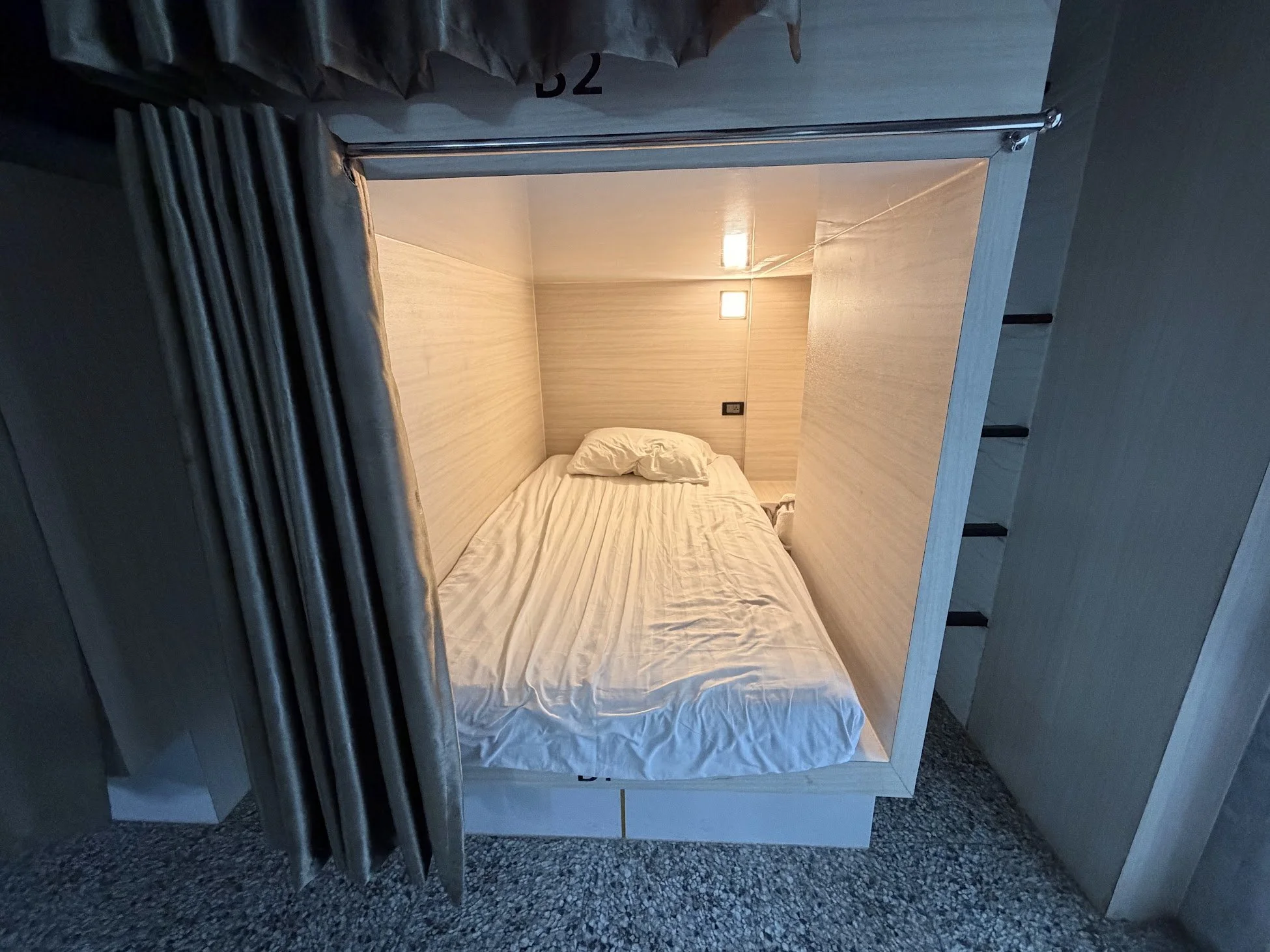 A large capsule hostel bed seen from outside the bed, with a cubby for luggage, light, plug and curtain for privacy, at White Jail Hostel in Koh Tao, Thailand.