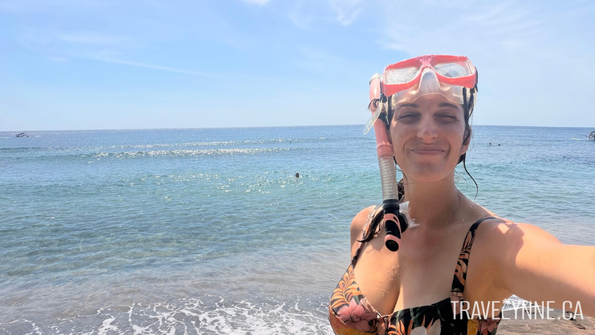 A woman wearing a bathing suit takes a selfie with the ocean in Amed, Bali while wearing a snorkel and mask on her head