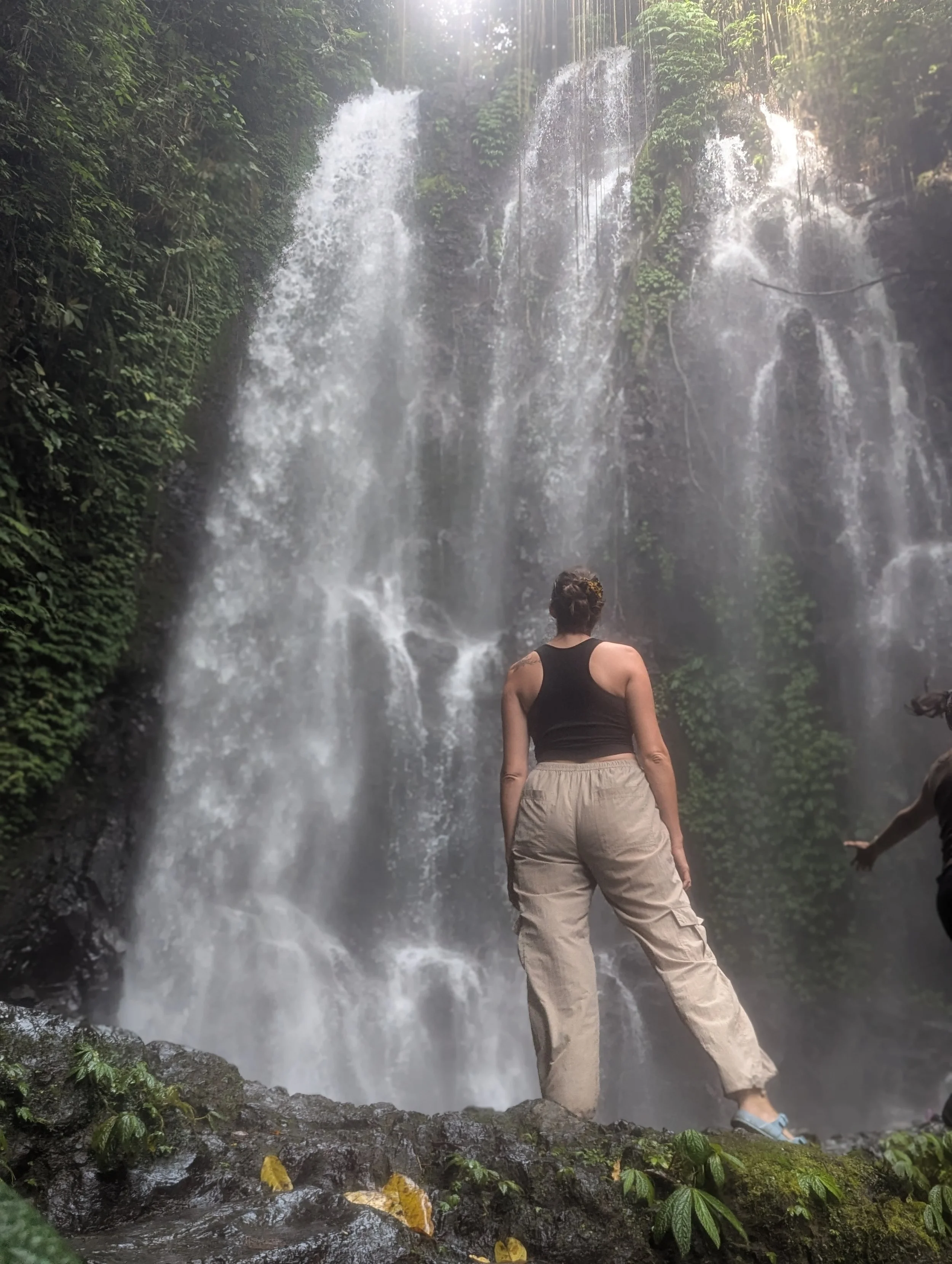 A woman stands facing a waterfall in a misty aired forest