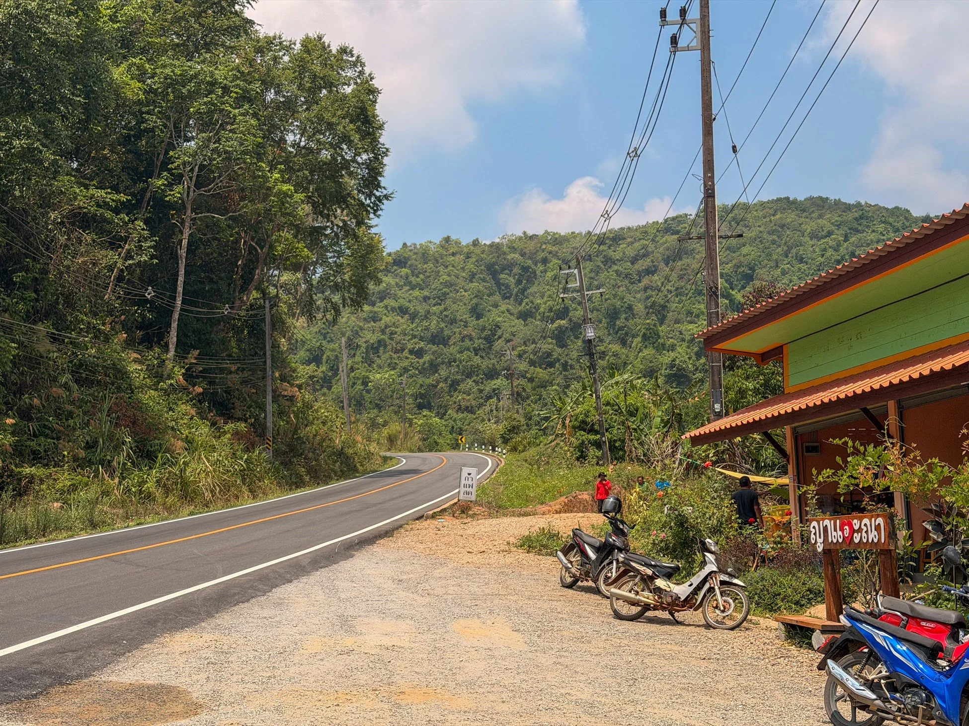 A paved road curves through a forest on the Mae Hong Son Loop with a cafe next to it with scooters parked out front