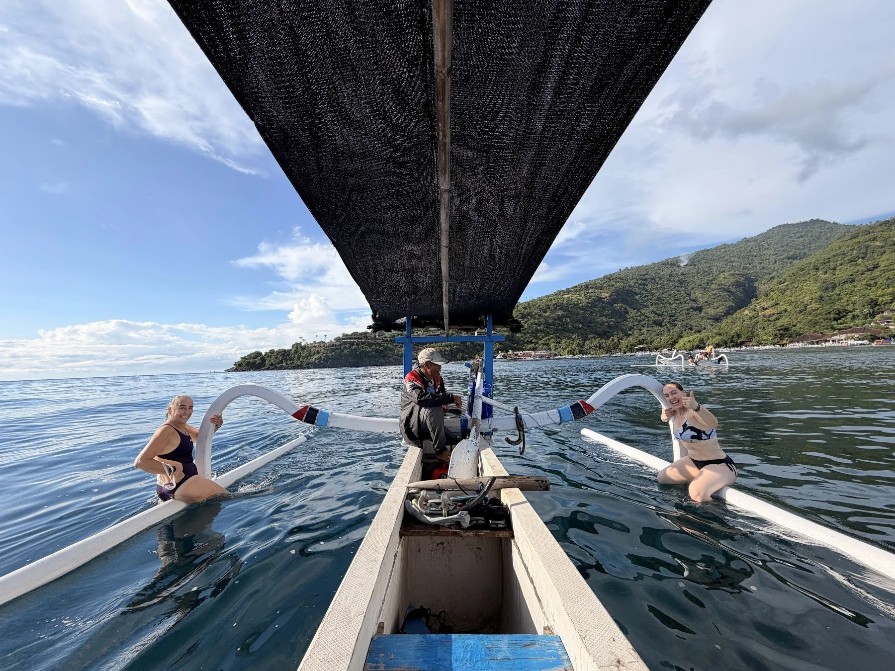 A fisherman sits in his traditional fishing boat on the sea with Amed visible on the coast, and two women sit on either side of the boat giving thumbs up