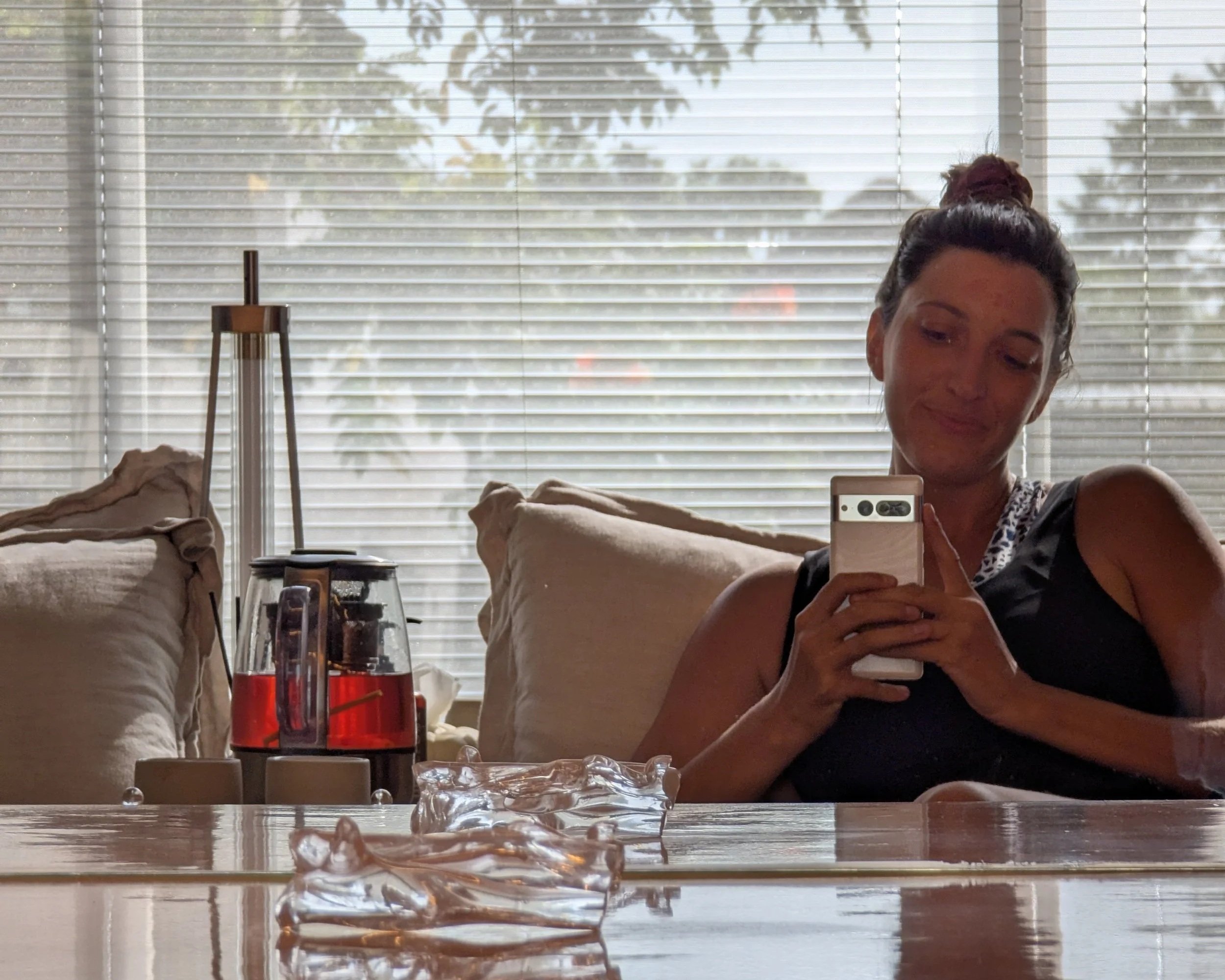 A woman takes a selfie in the mirror at a spa, with trees outside behind her and a pot of rosella tea next to her. She has a high bun and is holding a white phone.