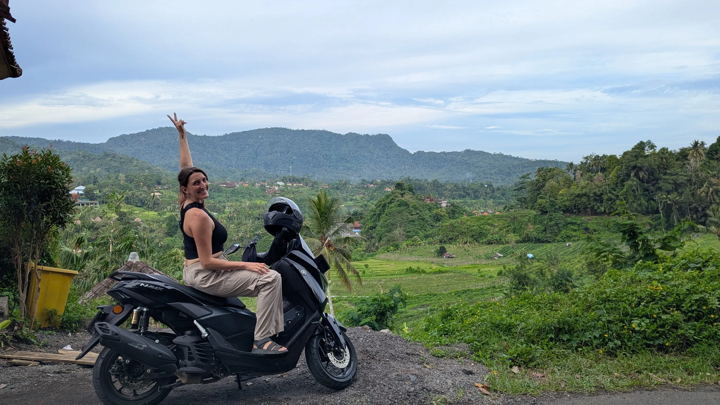 A woman makes a peace sign in the air while sitting on an NMAX scooter overlooking a valley with farmland in Sidemen, Bali.
