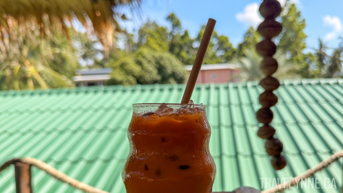 A close up of an orange Thai tea in a curvy glass with a bamboo straw,  with trees and a green roof in the background.