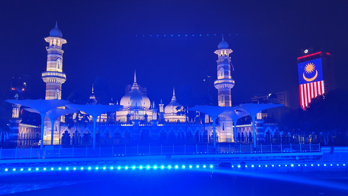 Masjid Jamek, a mosque in Chinatown Kuala Lumpur, is lit up in blue at night, with a Malaysian flag hanging next to it, and the river of life in the forefront