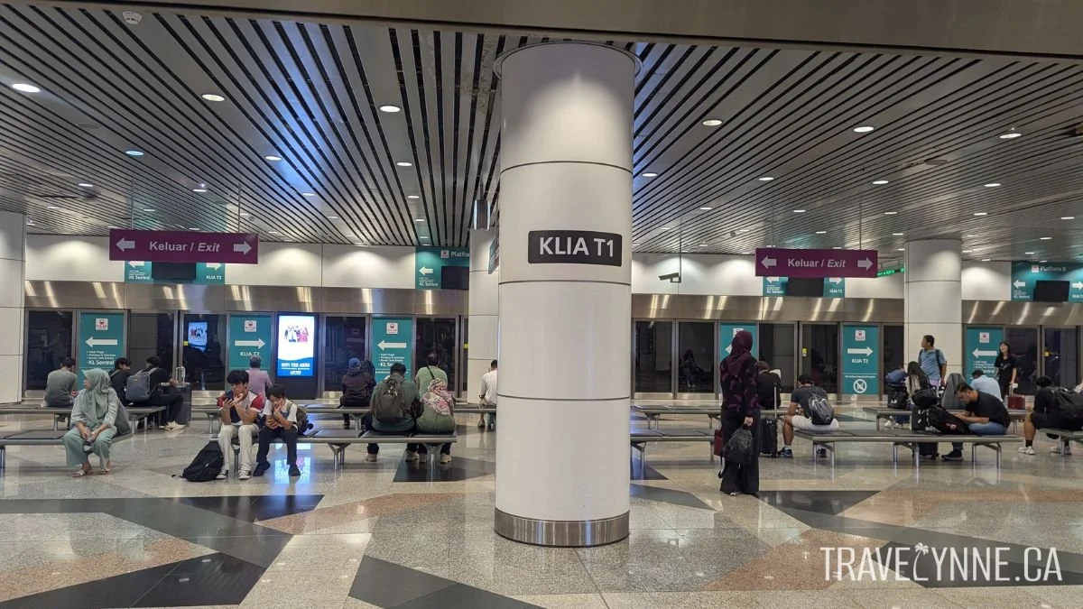 A "KLIA T1" sign on a post in the middle of the waiting area for the KLIA Ekspres train in the KLIA airport, with several passengers waitinf on benches.