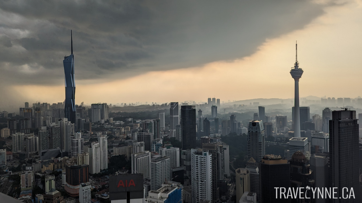 The Kuala Lumpur skyline seen after rain, with dramatic grey clouds opening to a dusty orange sky, a long urban skyline featuring Merdeka Tower and KL Tower