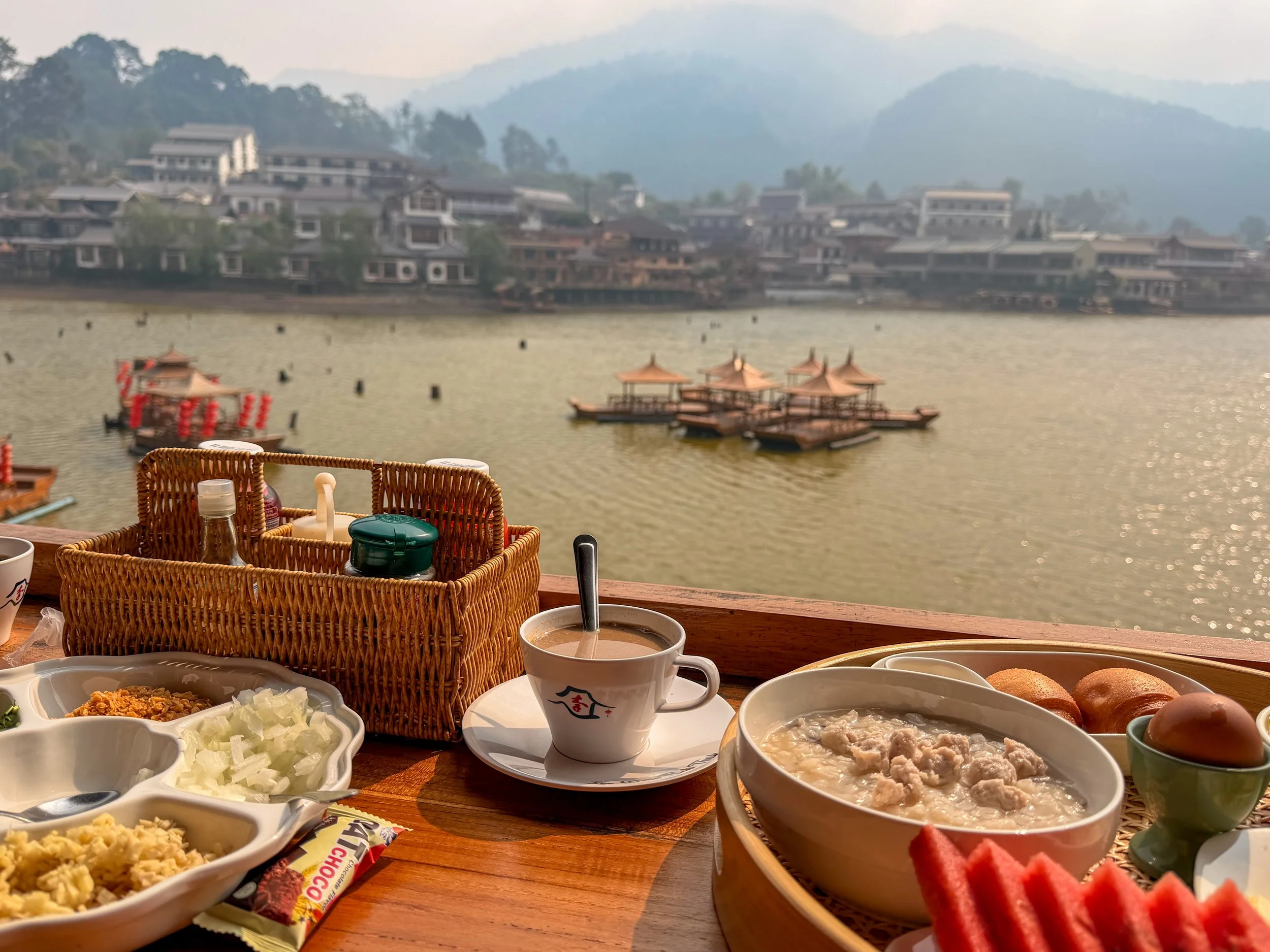 A Chinese-Thai breakfast of savory porridge, buns, noodles, watermelon and coffee in front of a lake with Chinese-style wooden boats in the background at Lee Wine restaurant in Bang Rak Thai, on the  Mae Hong Son Loop in Thailand.