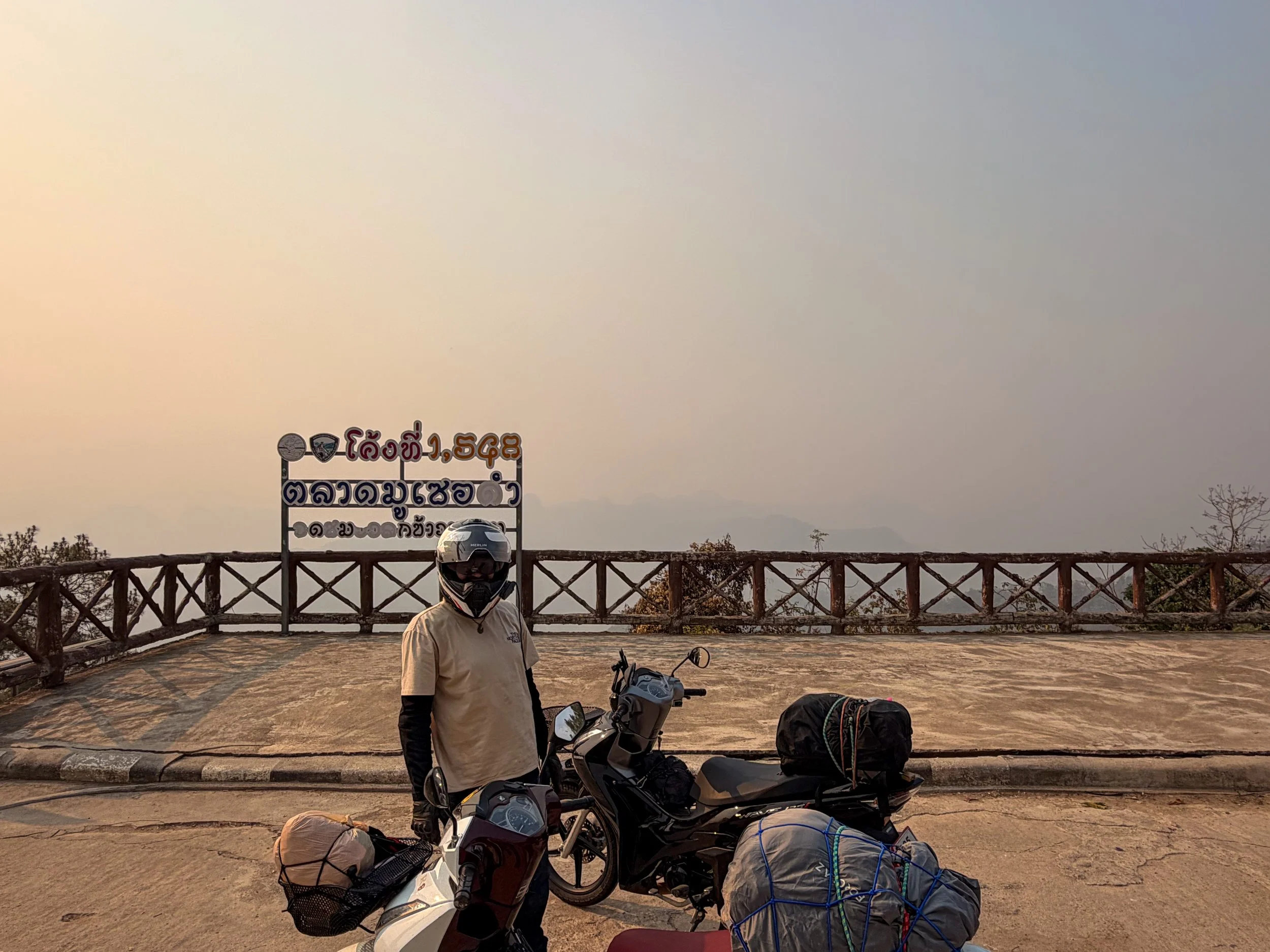 A rider stands with his scooter on the Mae Hong Son Loop, with the background washed out by thick haze from the smoky season in Northern Thailand.