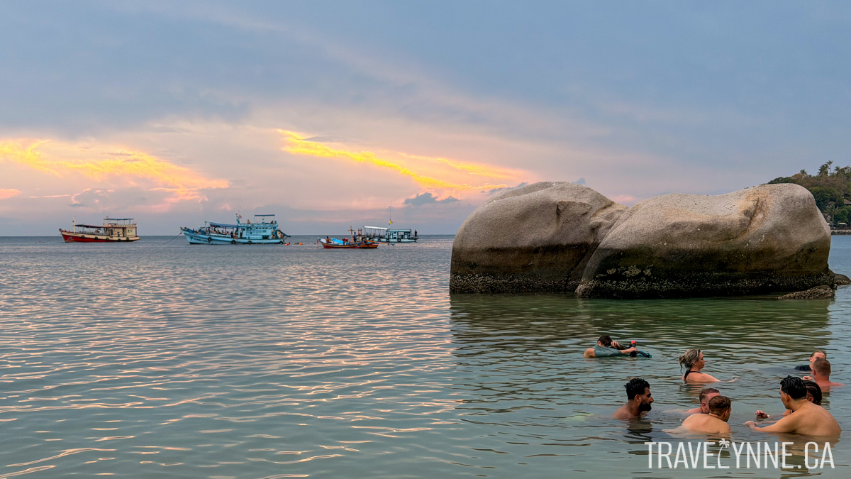 Swimmers bathe next to giant rocks in the sea at Freedom Beach, Koh Tao, while colorful boats float behind and the sun sets