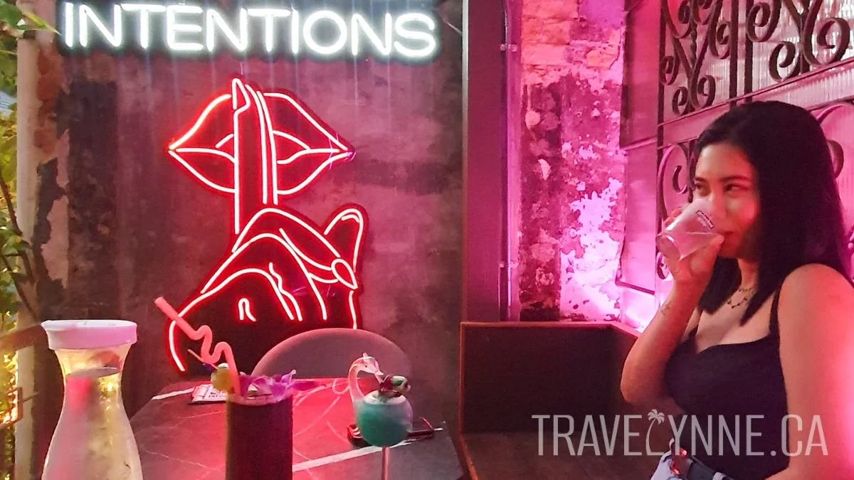 A beautiful woman with dark hair has a drink on an outdoor terrace at GSting bar in Chinatown, KL, with a background of neon lights including a large light of a finger shushing lips and the word "INTENTIONS".