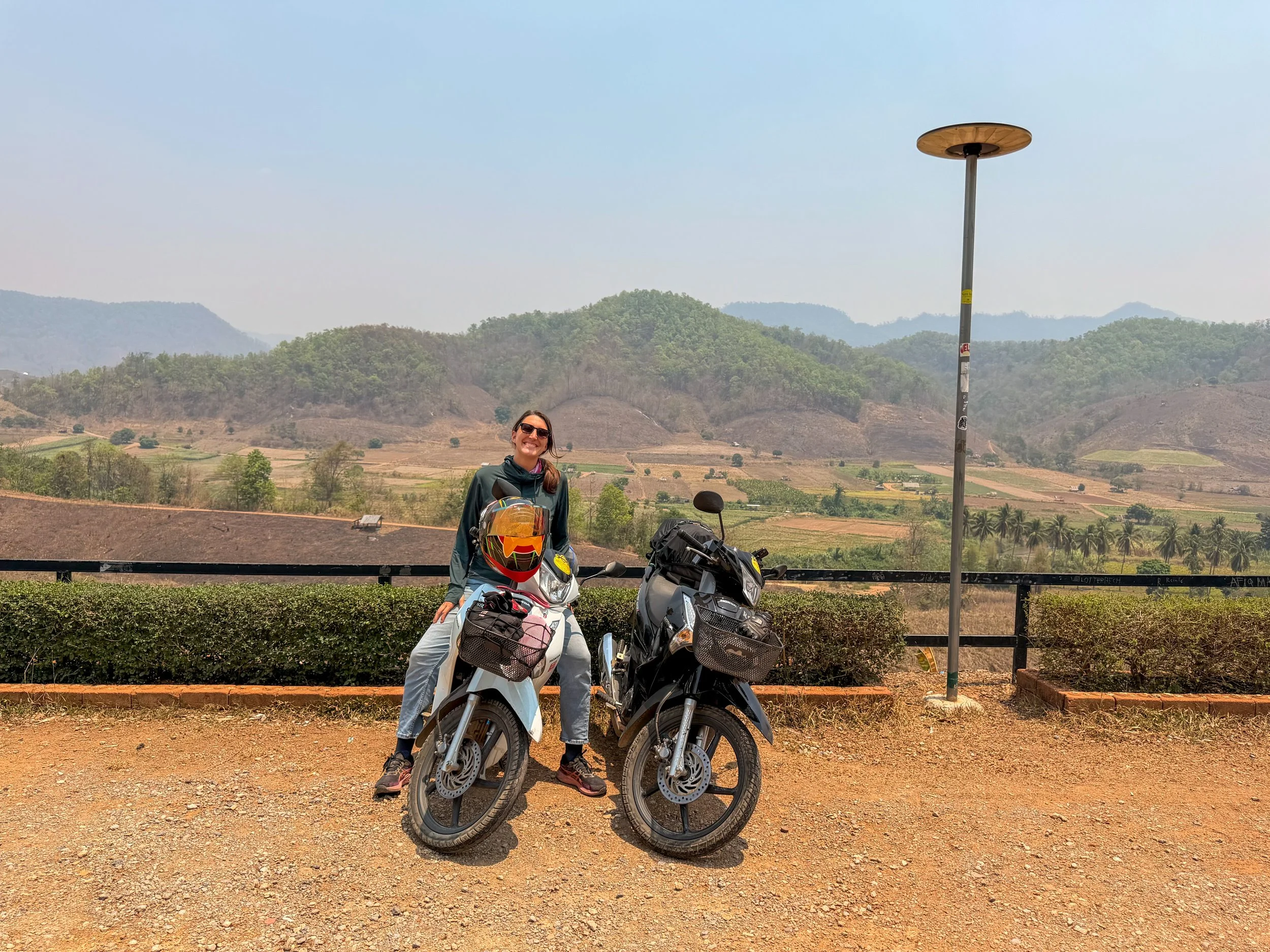 Lynne sits on one of two semi-automatic scooters with a hilly backdrop on the Mae Hong Son Loop in Northern Thailand.