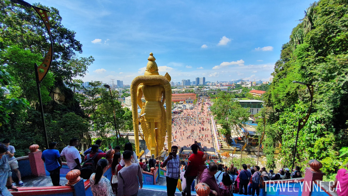 The view from mid-way up the stairs at Batu Caves temple, with the back of a giant golden Lord Murugan statue, many people coming and going on the stairs, and the city skyline in the distance.
