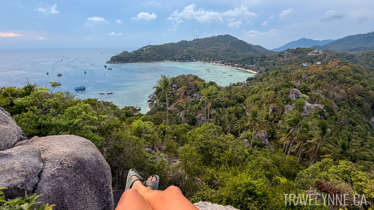 Lynne's legs dangle off a cliff overlooking green palmy jungles and a blue and turquoise bay at John-Suwan Viewpoint in Koh Tao.