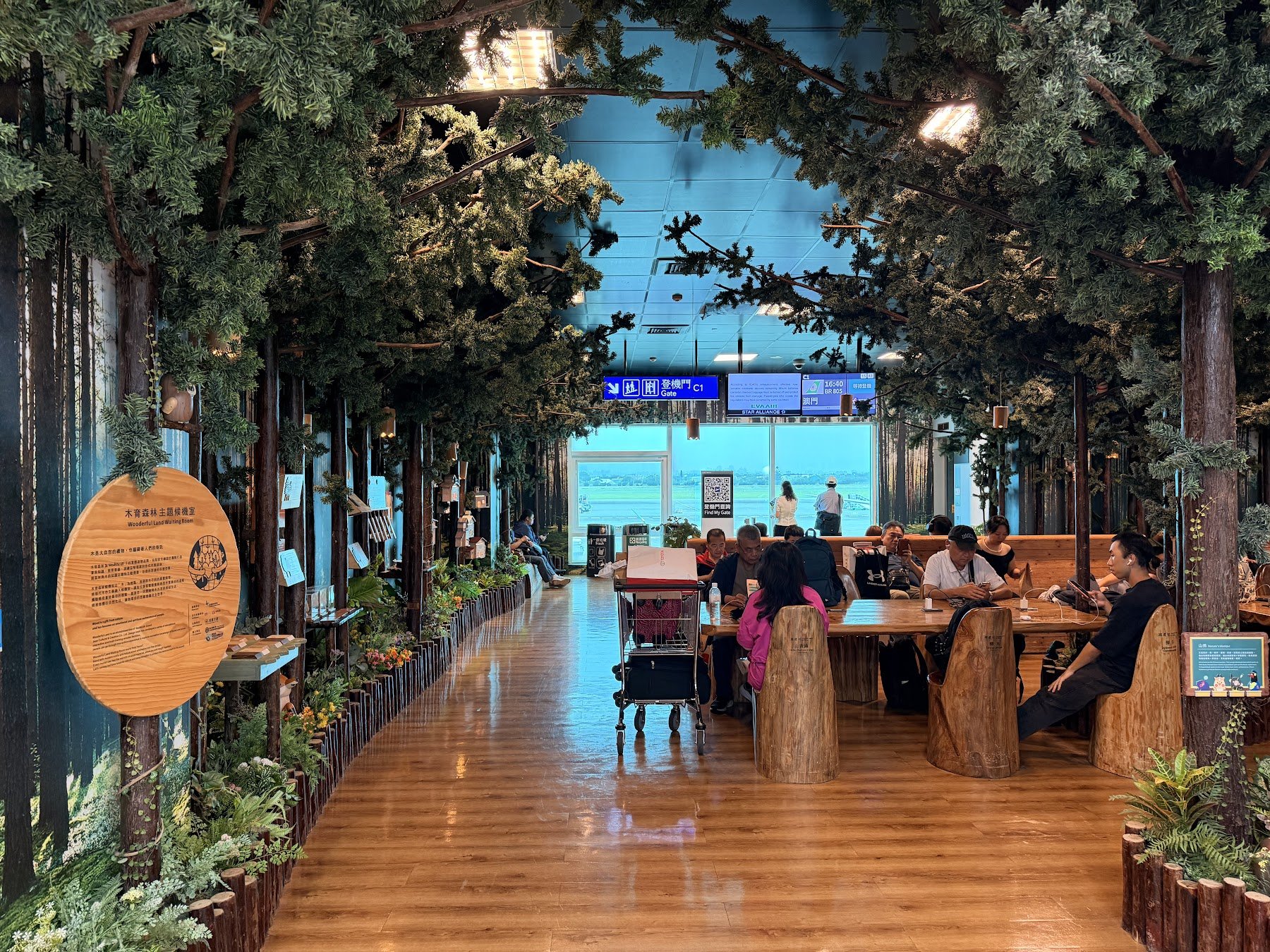 A workspace located in Taiwan Taoyuan International Airport Terminal 2, with a forest theme there are fake trees and shrubs, wooden floors, wooden tables sand chairs made of logs. People are charging devices and working from the tables.