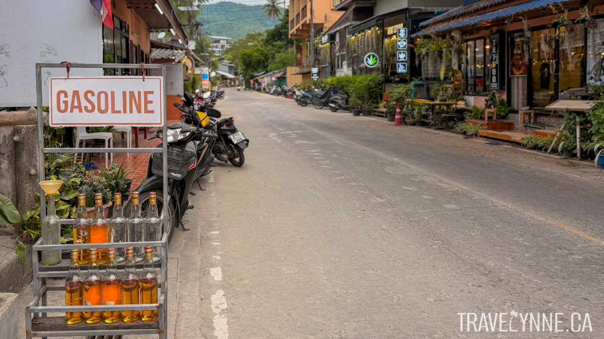 A gasoline rack on a road in Koh Tao, Thailand, with recycled glass bottles housing gas, mopeds parked along the road, and restaurants and cannabis stores on the street.