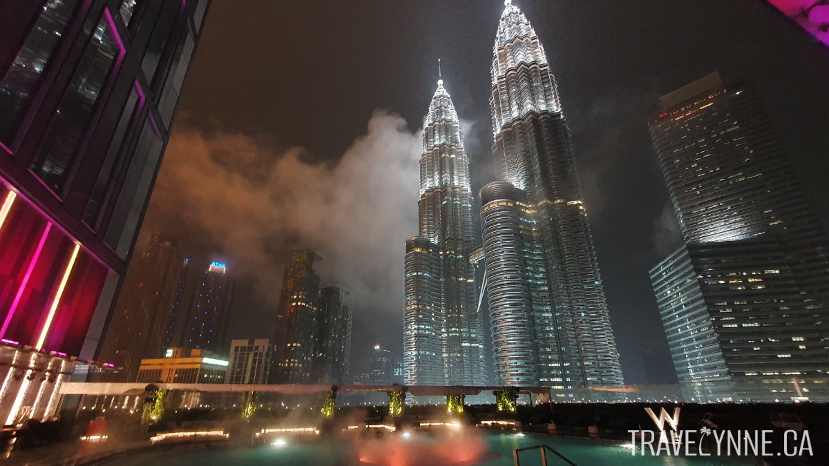 A night sky view of the Petronas Twin towers from the Wet Deck bar in Kuala Lumpur, with the twin towers lit up and a cloudy night sky reflecting on a pool
