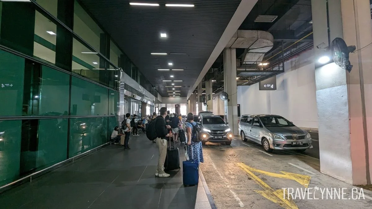 Travellers wait for their rideshare vehicles outside of the pickup point in KLIA2 airport terminal, with silver cars lined up in the underground parking space