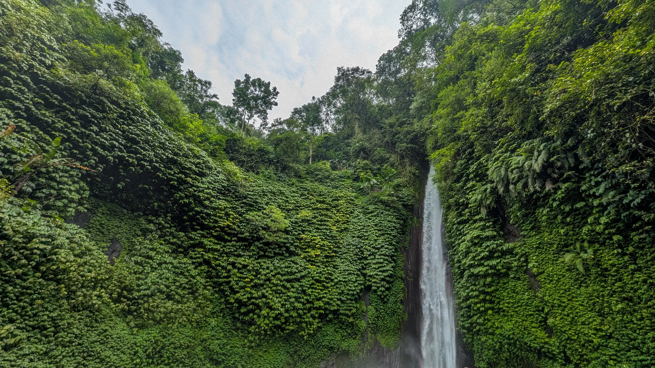 The sky peeks out above a wall of bright green foliage with a narrow but strong stream of water from Red Coral Waterfall in Munduk, Bali