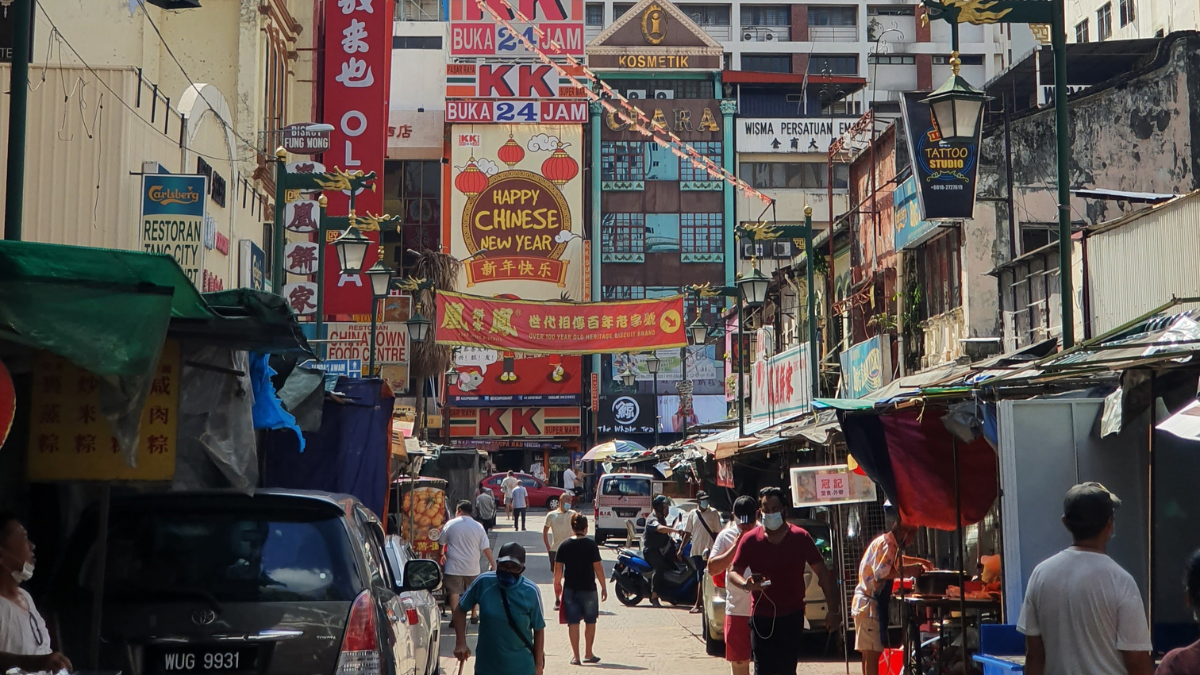 A Chinatown market street in Kuala Lumpur, with Chinese and Malay signs hanging from buildings, people walking about carrying bags, and cars and scooters getting by in the narrow road