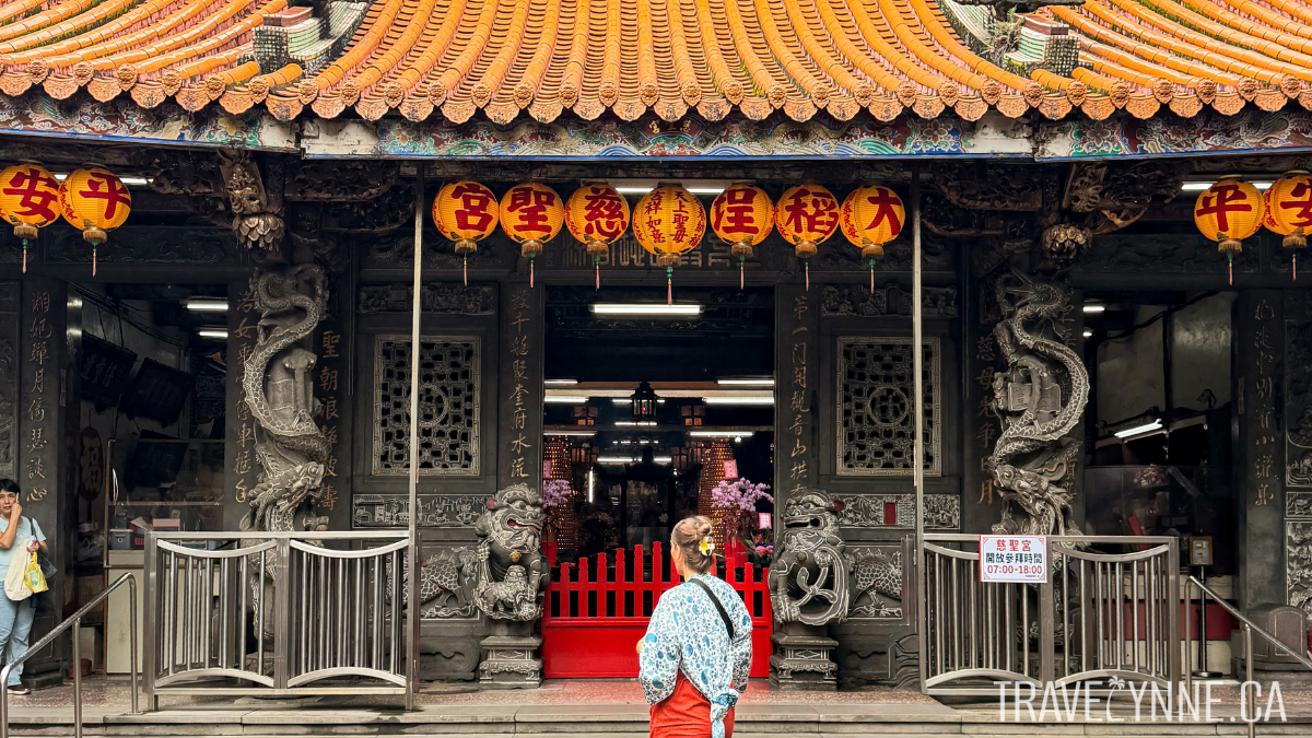 Wearing a blue and white floral kimono, Lynne faces away from the camera admiring a temple in Taipei, with a traditional temple roof and lanterns overhead