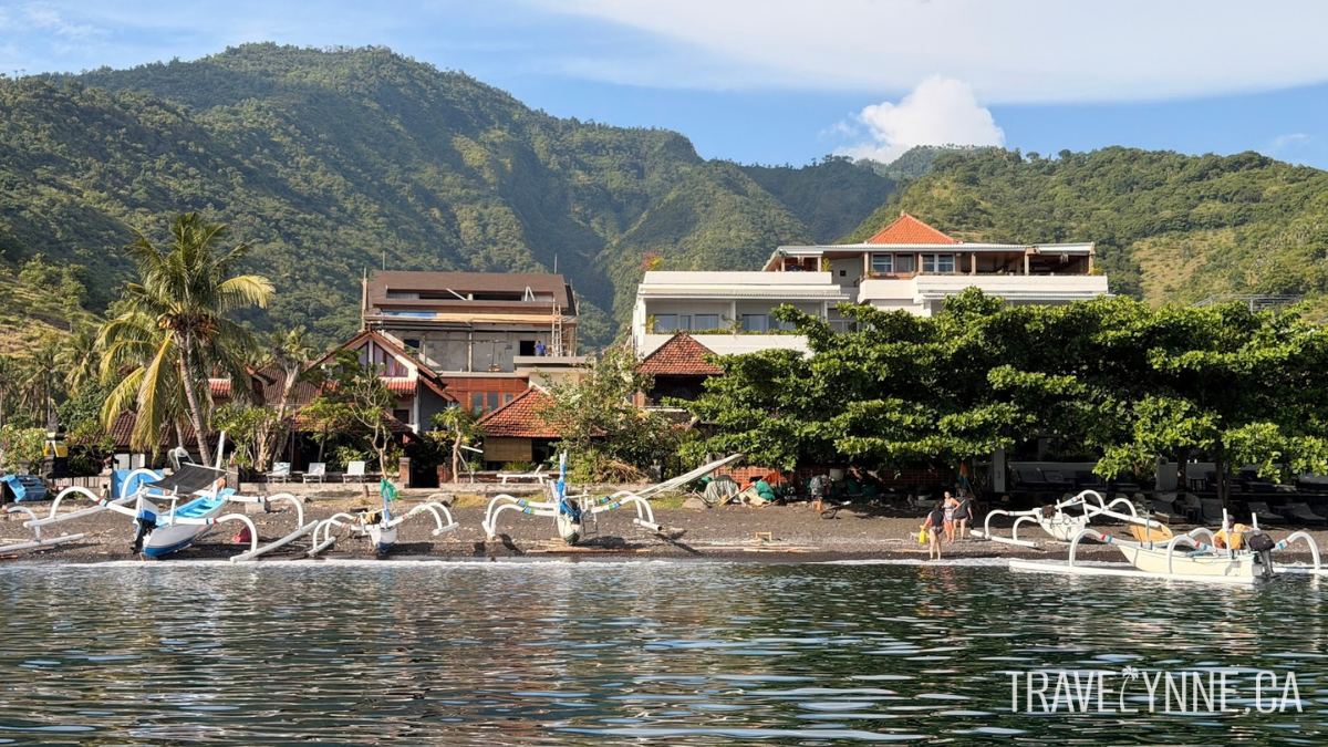 A view of the coastline in Amed, Bali, with green hills, low rise hotels, palm trees, fishing boats, and a black sand beach