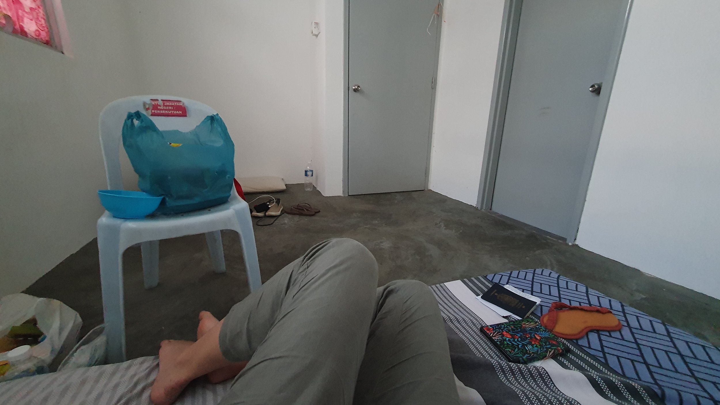 From the POV of the photographer, you see her legs on a small mattress with her passport, eye mask and ereader, on a cement floor with a plastic chair and bag of food, in a quarantine room.