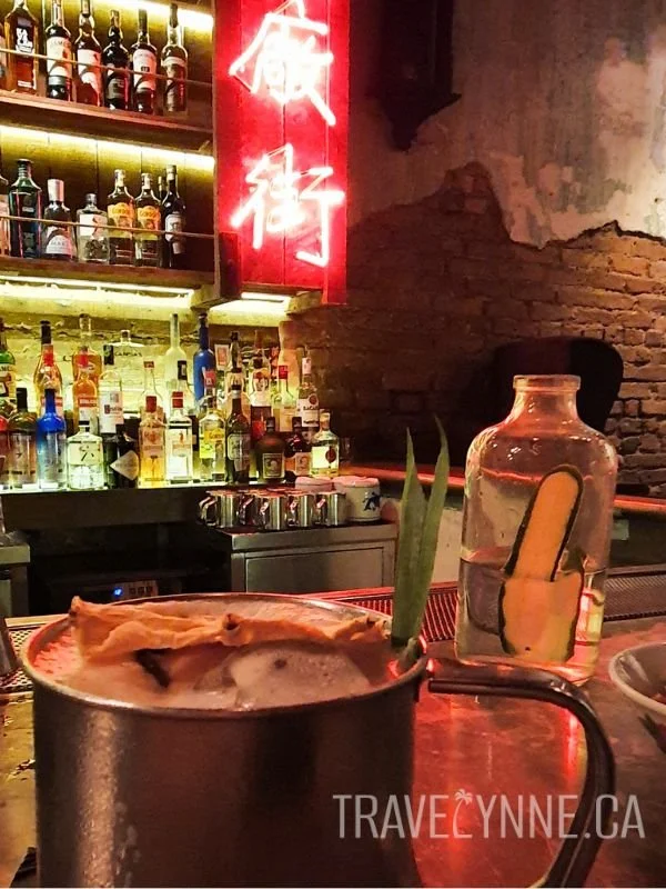 A fruity drink adorned with dried pineapple stands in the foreground of a moody backlit bar with Chinese neon signage.