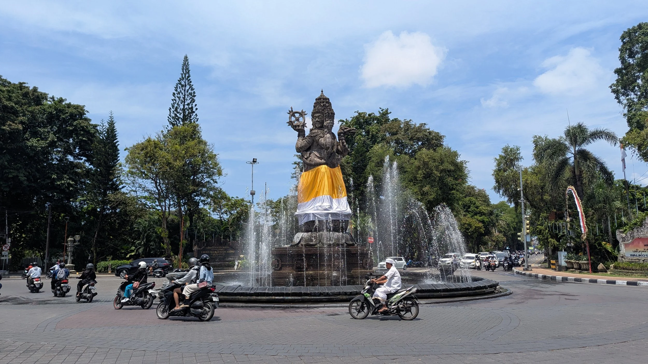 Scooter drivers whizz past an ornate roundabout in Denpasar, Bali, Indonesia, with a Hindu statue and fountain at its centre and trees in the background