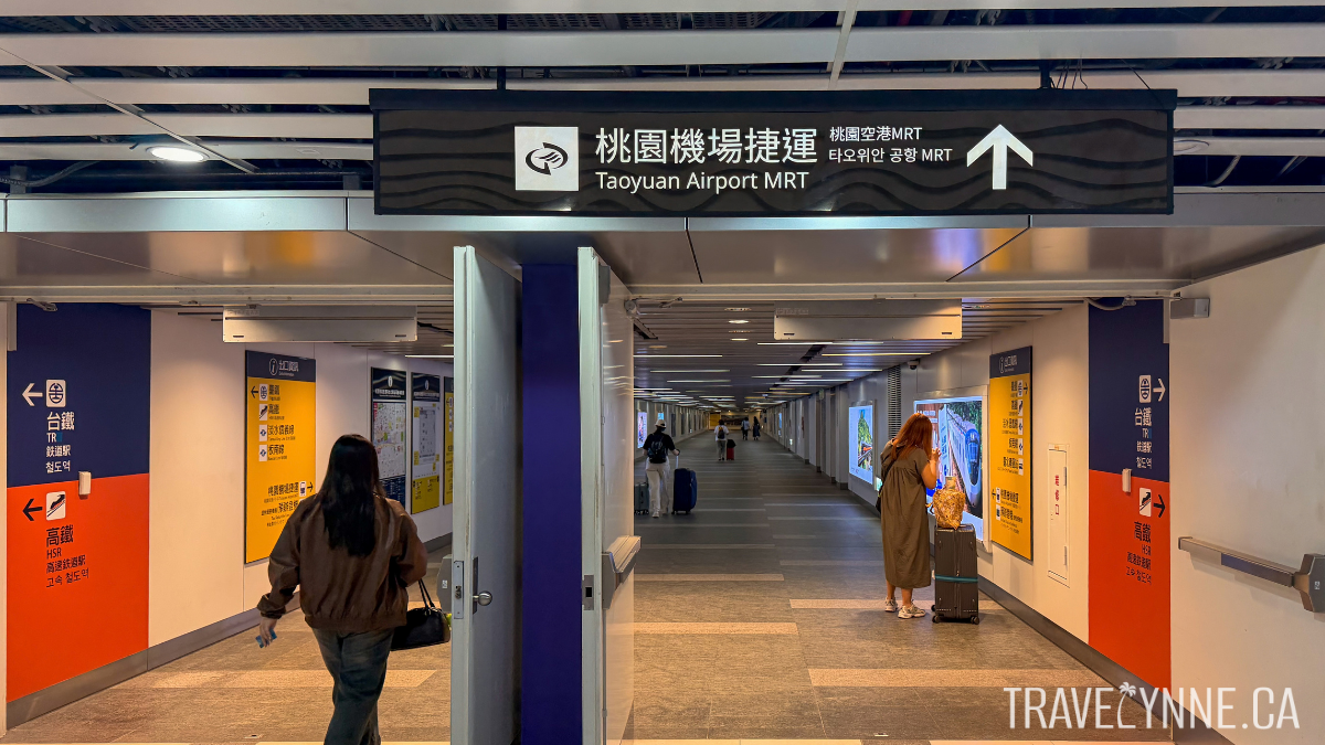 Overhead signage shows the direction to Taoyuan Airport MRT from Taipei Main Station, with a long hallway ahead and a few travelers walking with luggage