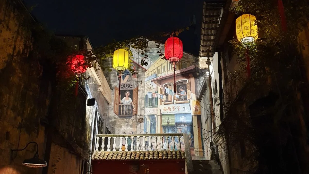 At night in the alleyway behind Concubine bar in Chinatown, red and yellow lanterns glow, and a mural of an Old Malaysia scene is lit up.