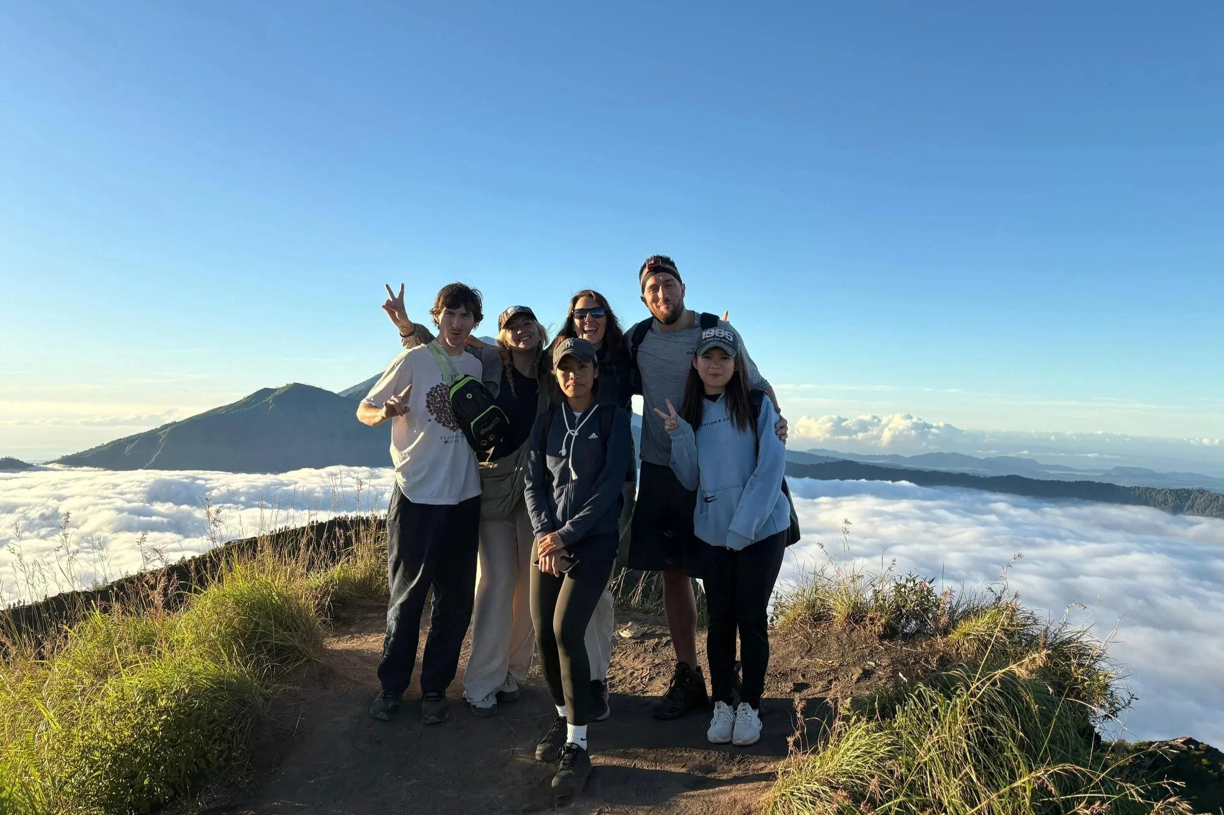 A group of American and Canadian tourists  stand proudly at the top of Mount Batur, Bali, with three female local guides