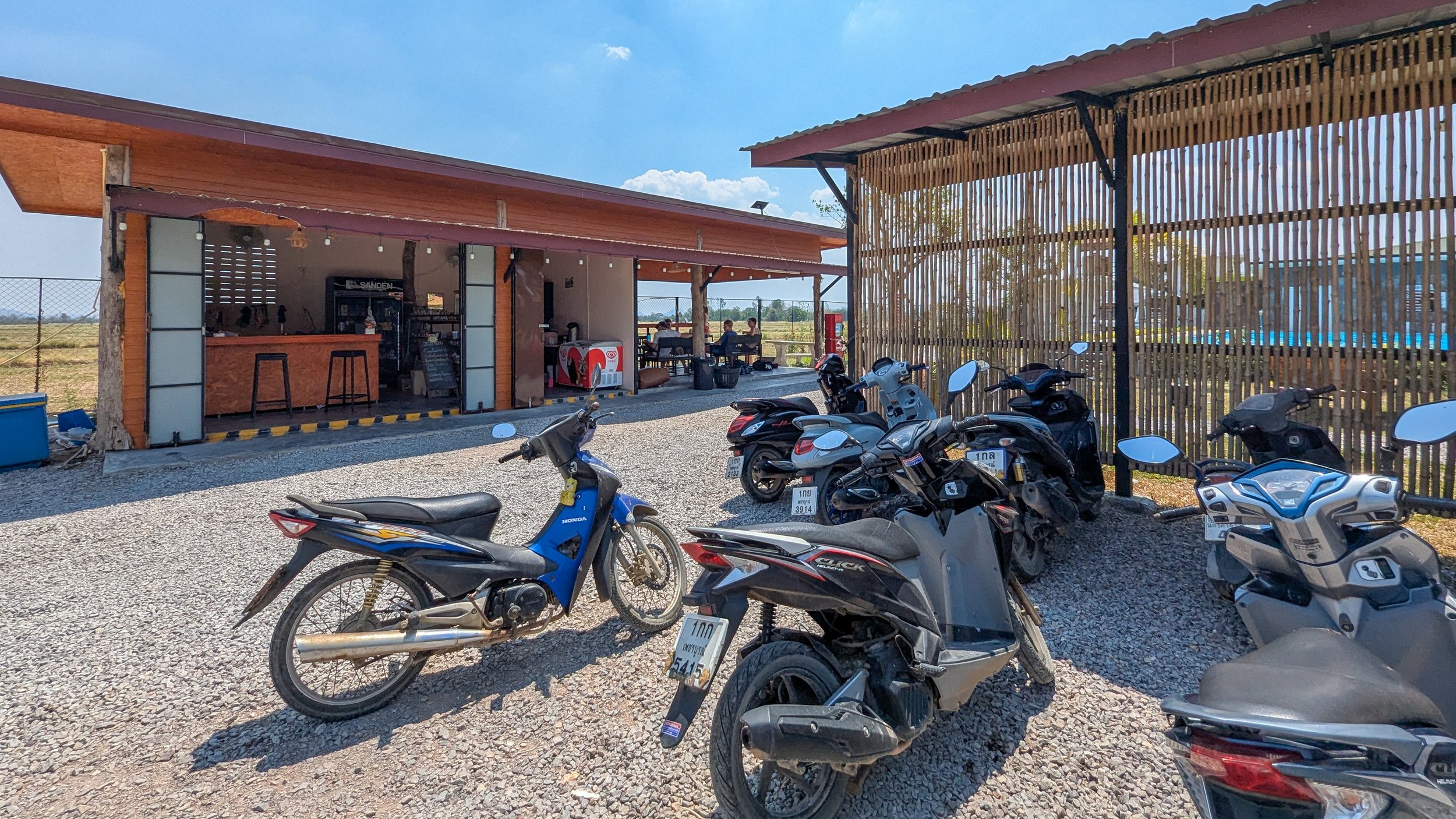 A variety of scooters are parked in a gravel parking space by a cafe in rural Thailand