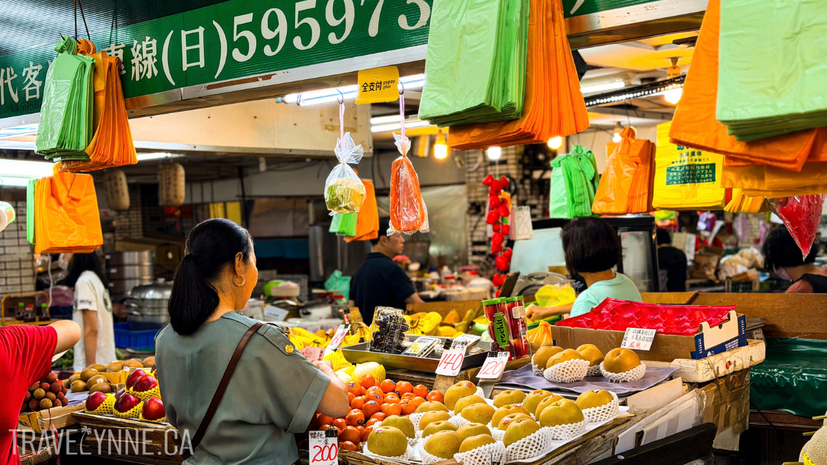 A woman looks at mandarins at a shop in a local market in Taipei, with lots of colourful fruits like apples and mangoes, and plastic bags hanging overhead