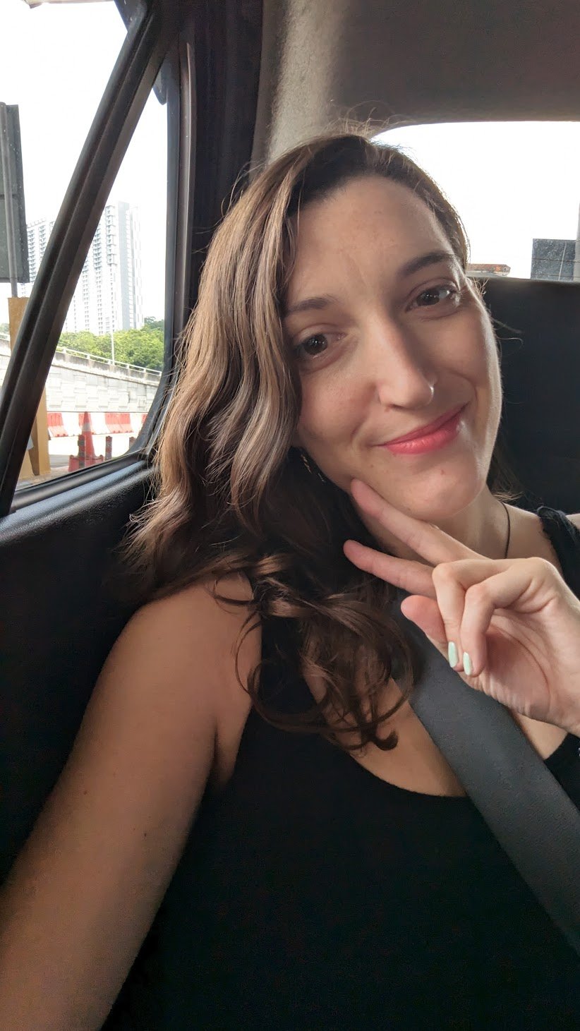 A dark haired woman makes a peace sign and takes a selfie sitting in the back seat of a car in a city, wearing a seatbelt