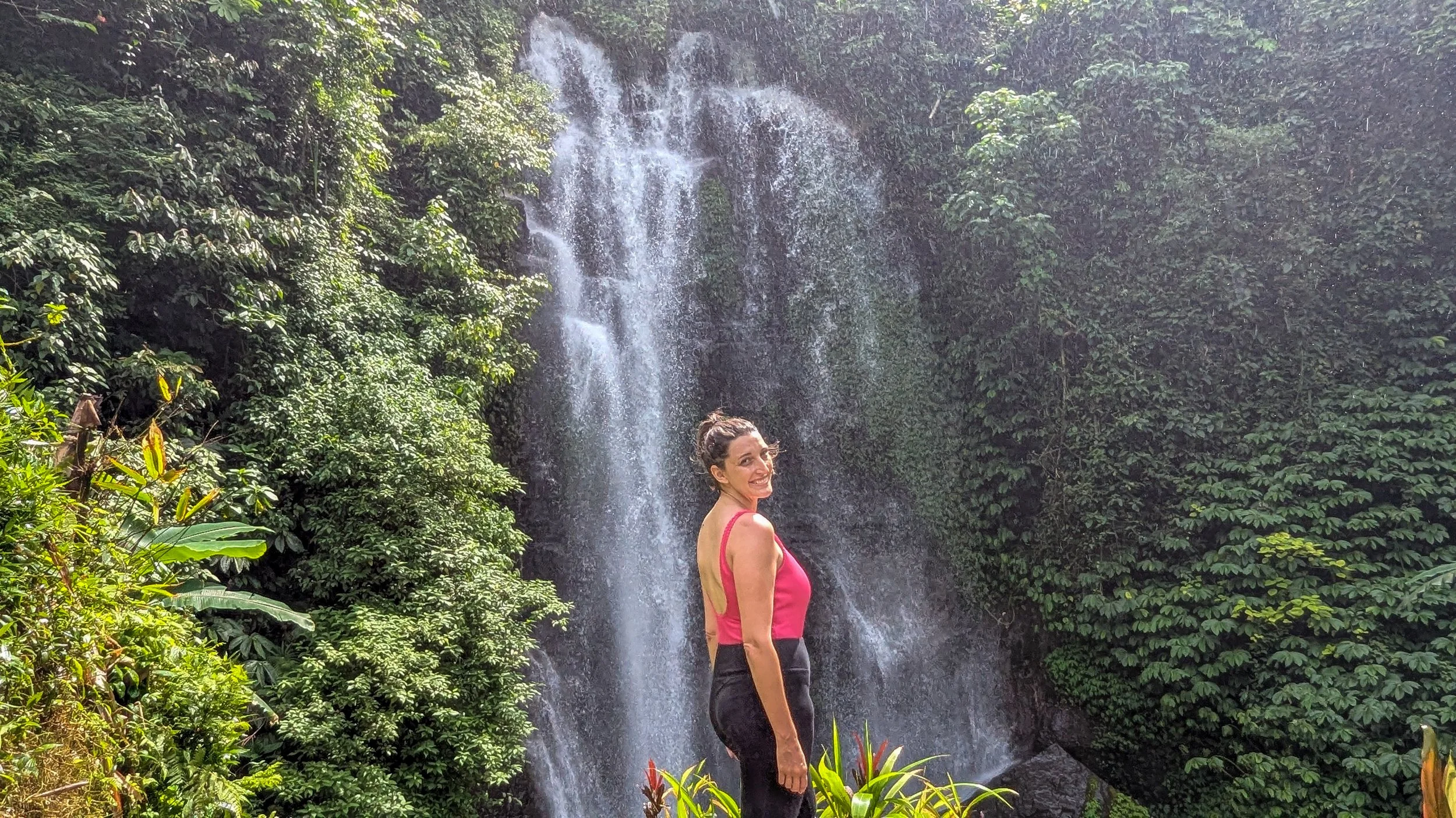 Woman in pink bathing suit and black leggings stands in front of Golden Valley waterfall in Munduk, Bali, surrounded by greenery