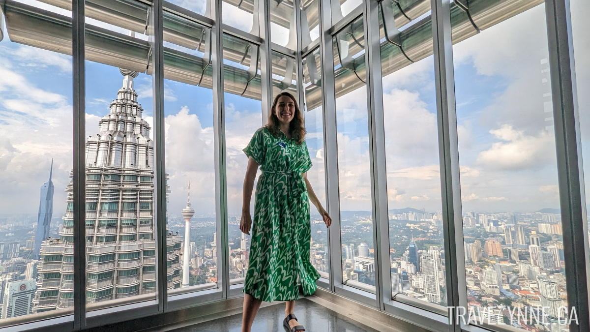 a woman wearing a bright green and white dress stands near windows on the observation deck of one of the Petronas Towers, with a clear view of the city skyline behind her, including the top of the other twin tower, the KL tower and the Merdeka Tower