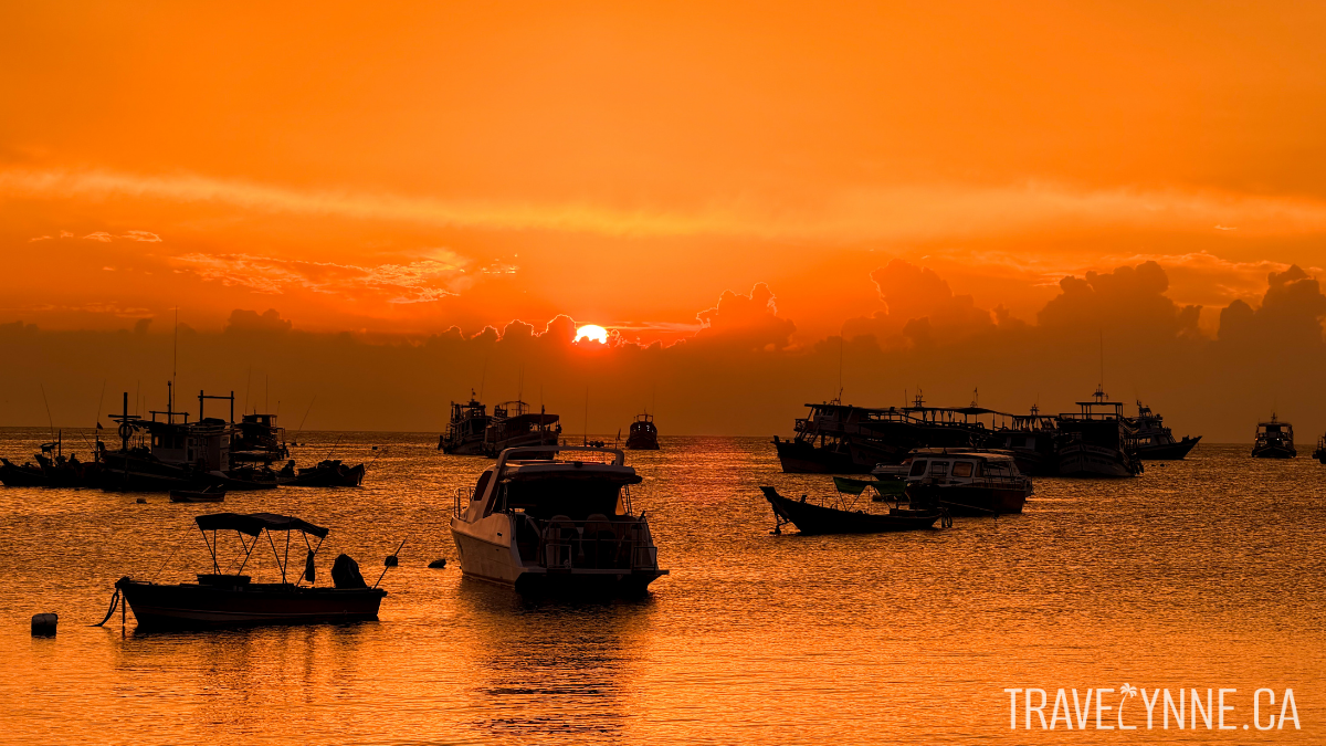 A dramatically orange sky is reflected on the sea in Sairee Beach, Koh Tao, with shadows of several boats in the horizon as the sun sets.