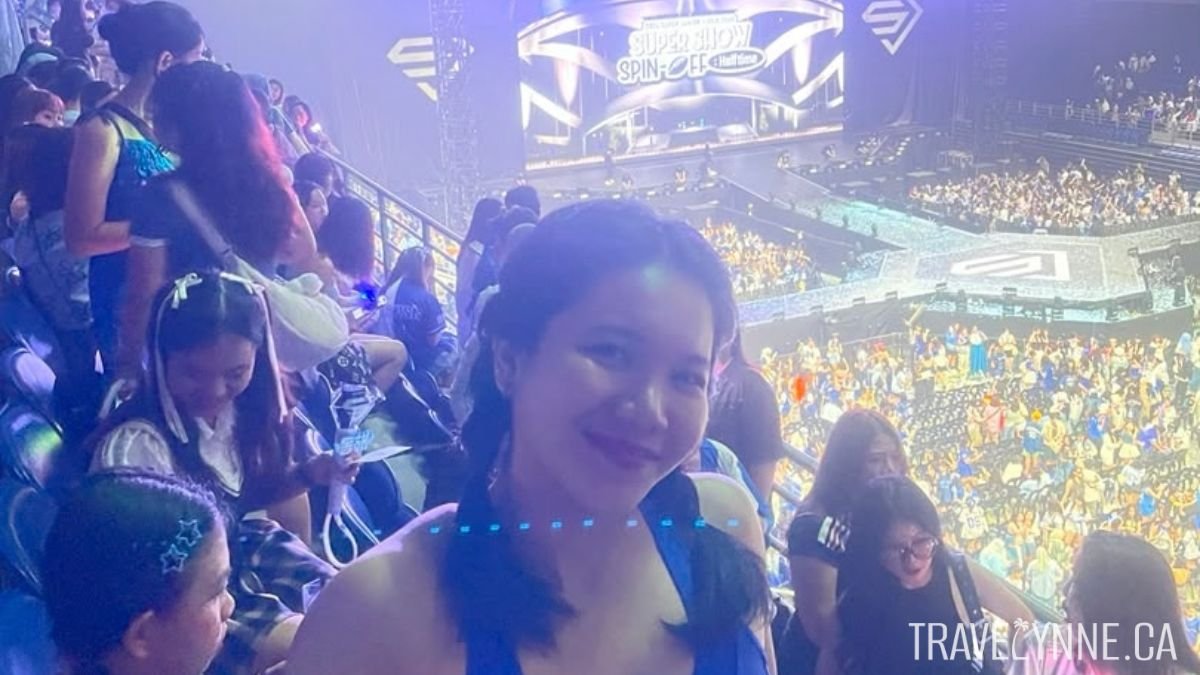 A woman smiles at the camera in Unifi Arena (Axiata Arena) before a Super Junior concert in KL, with fans getting to their seats in bleachers and on the floor. The screen says "SUPER SHOW SPIN OFF"