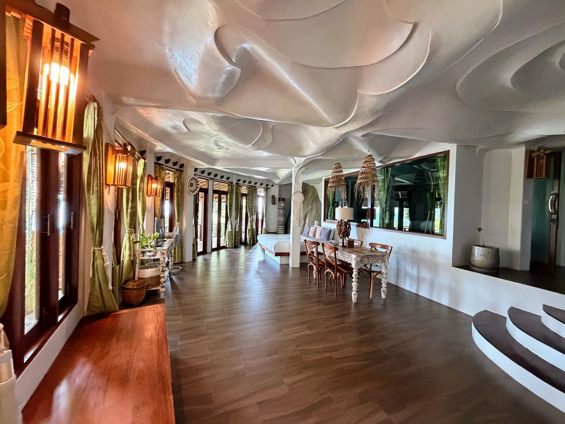 A unique family room interior is seen at a hotel in Koh Tao, Thailand, with a textured grey ceiling resembling waves, tropical style lamps, a large living area and bed in the back.